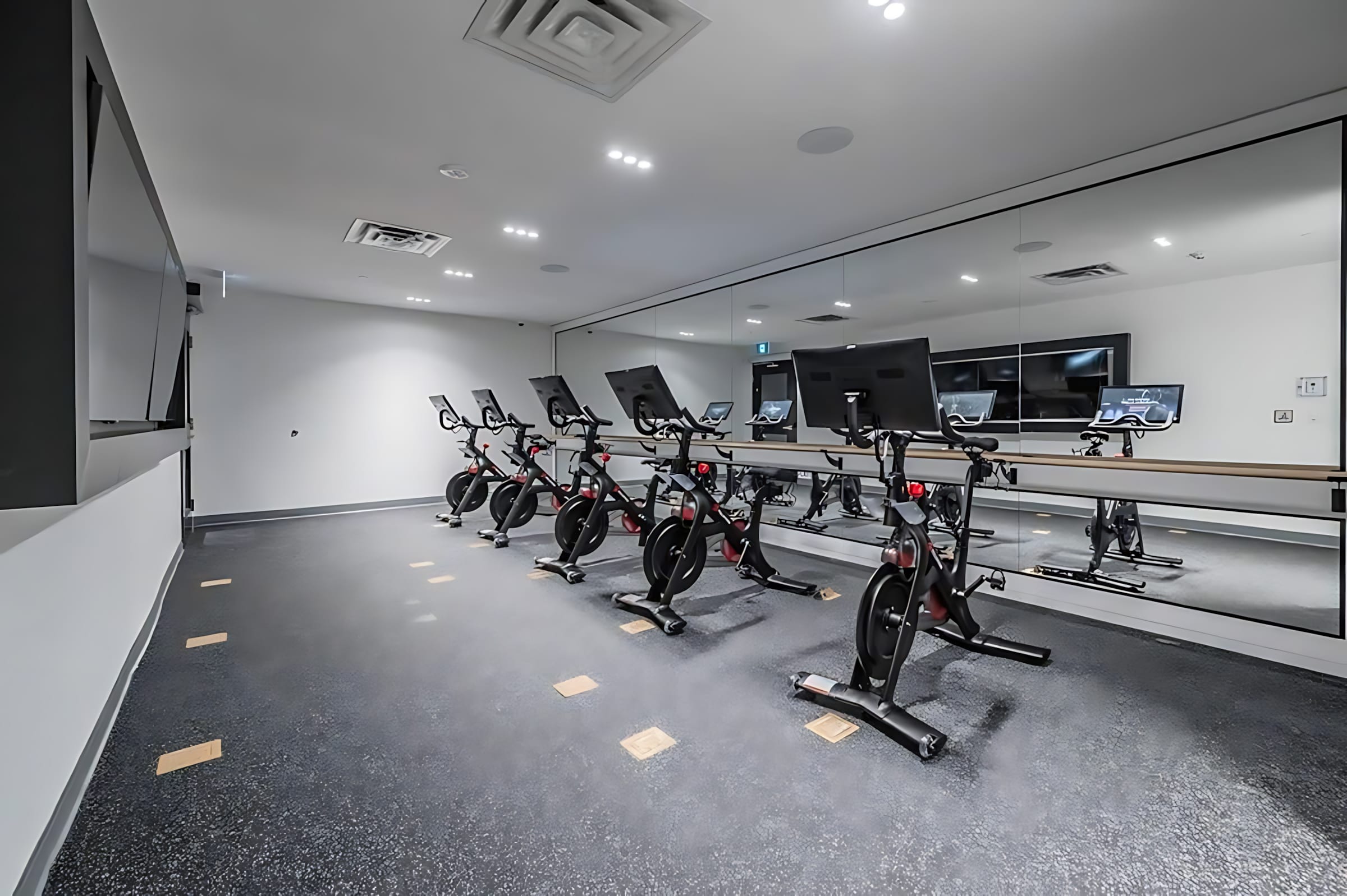 Dedicated cycling studio featuring a row of Peloton bikes facing a large mirrored wall, reflecting the room and screens. The dark rubber floor has light square markings, and the space is well-lit with recessed lights.