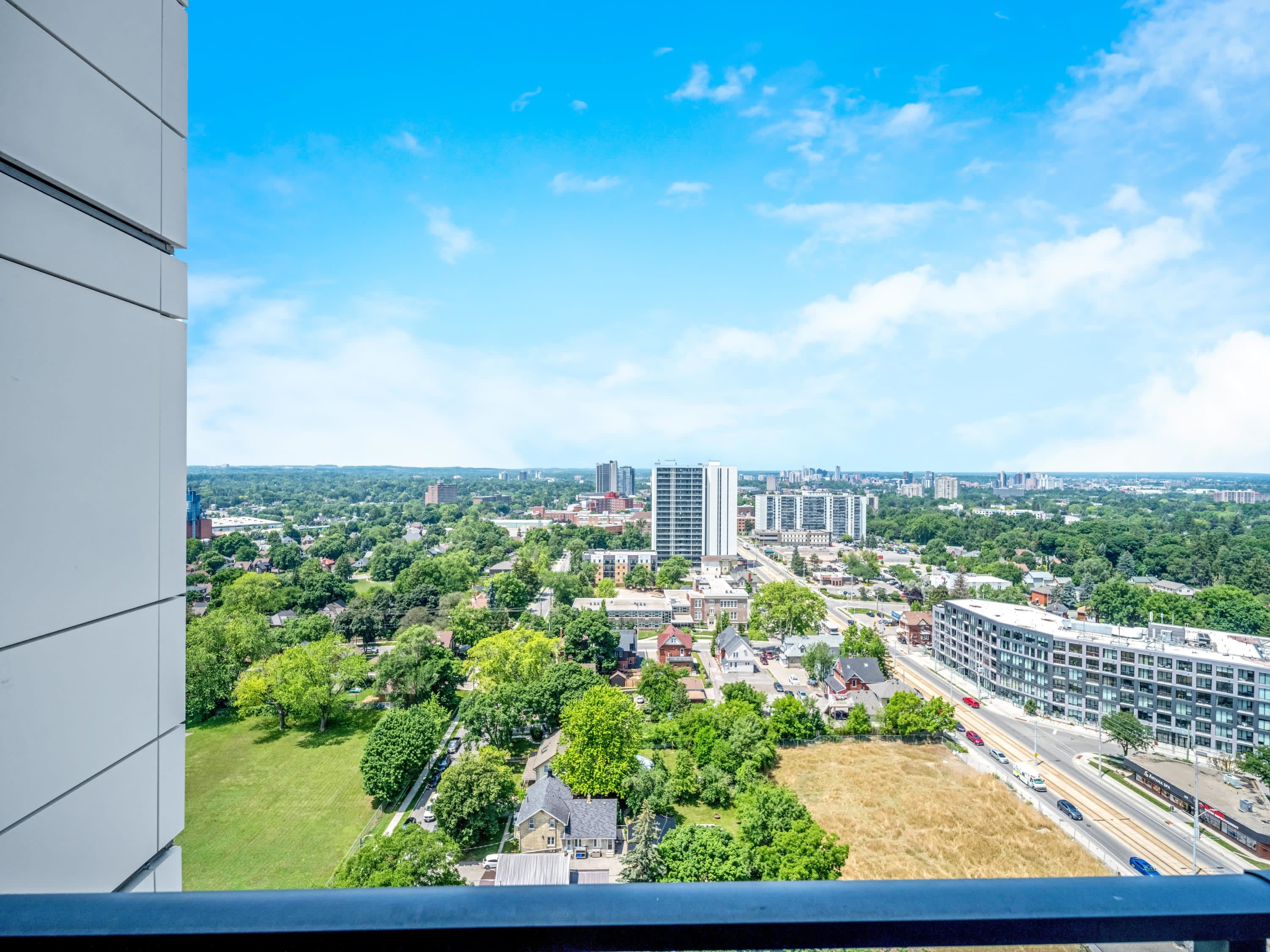 This image captures a stunning panoramic view from a high-rise balcony, overlooking a city with a mix of residential buildings, green spaces, and distant high-rises under a bright blue sky with scattered clouds.