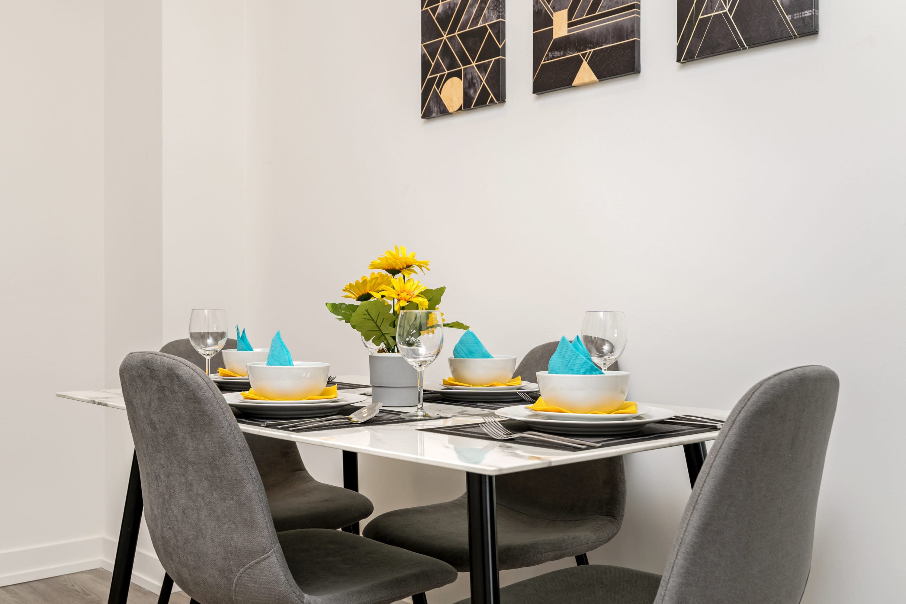 Close-up of a modern dining table set for four, featuring white bowls with blue napkins, wine glasses, and a yellow sunflower centerpiece. Abstract artwork is visible on the white wall.