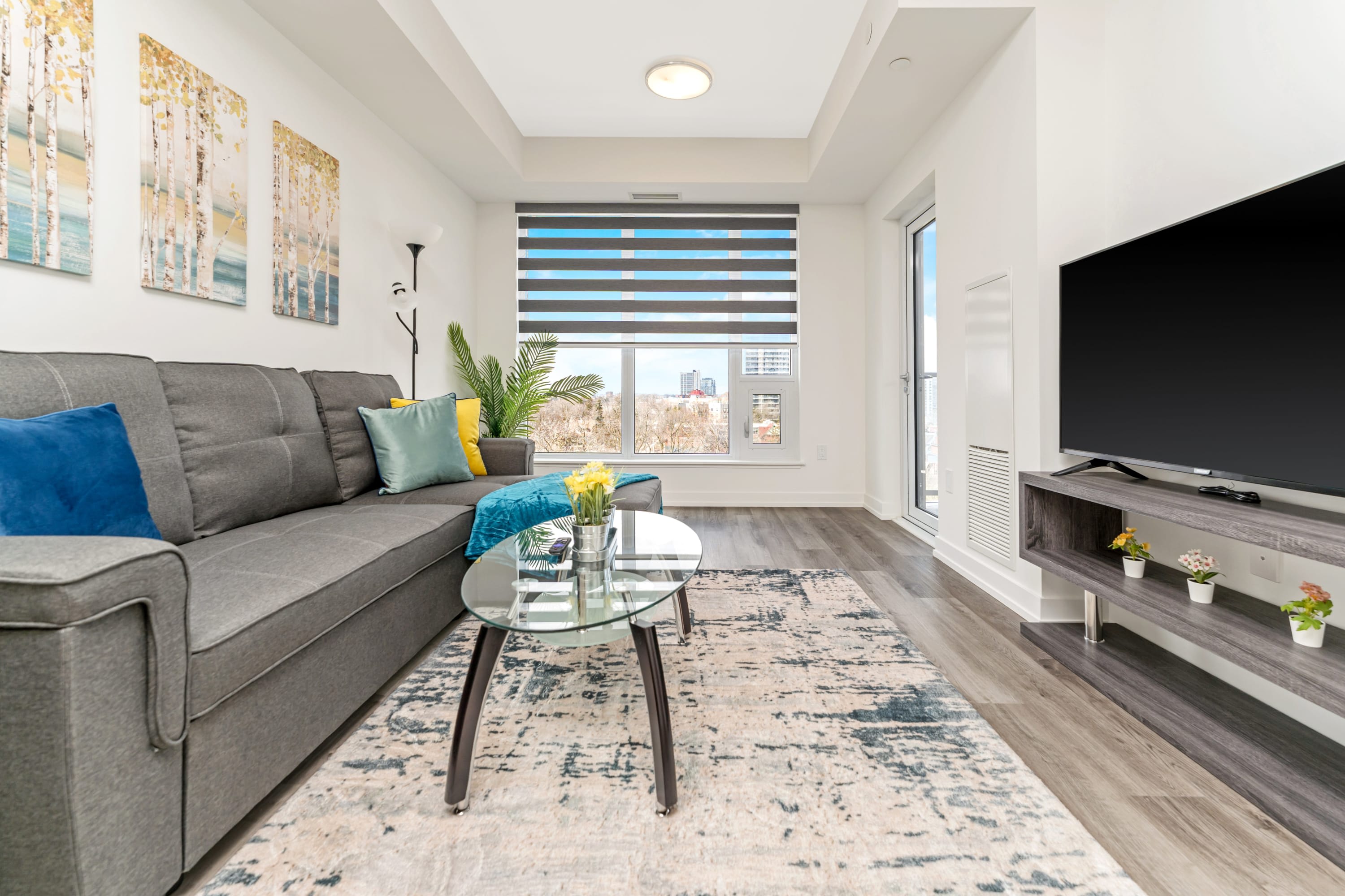A close-up view of the living room, highlighting the gray sectional sofa with colorful throw pillows and a teal blanket, a glass coffee table with flowers, and a large window with horizontal blinds offering city views. A floor lamp and a potted plant add to the decor.
