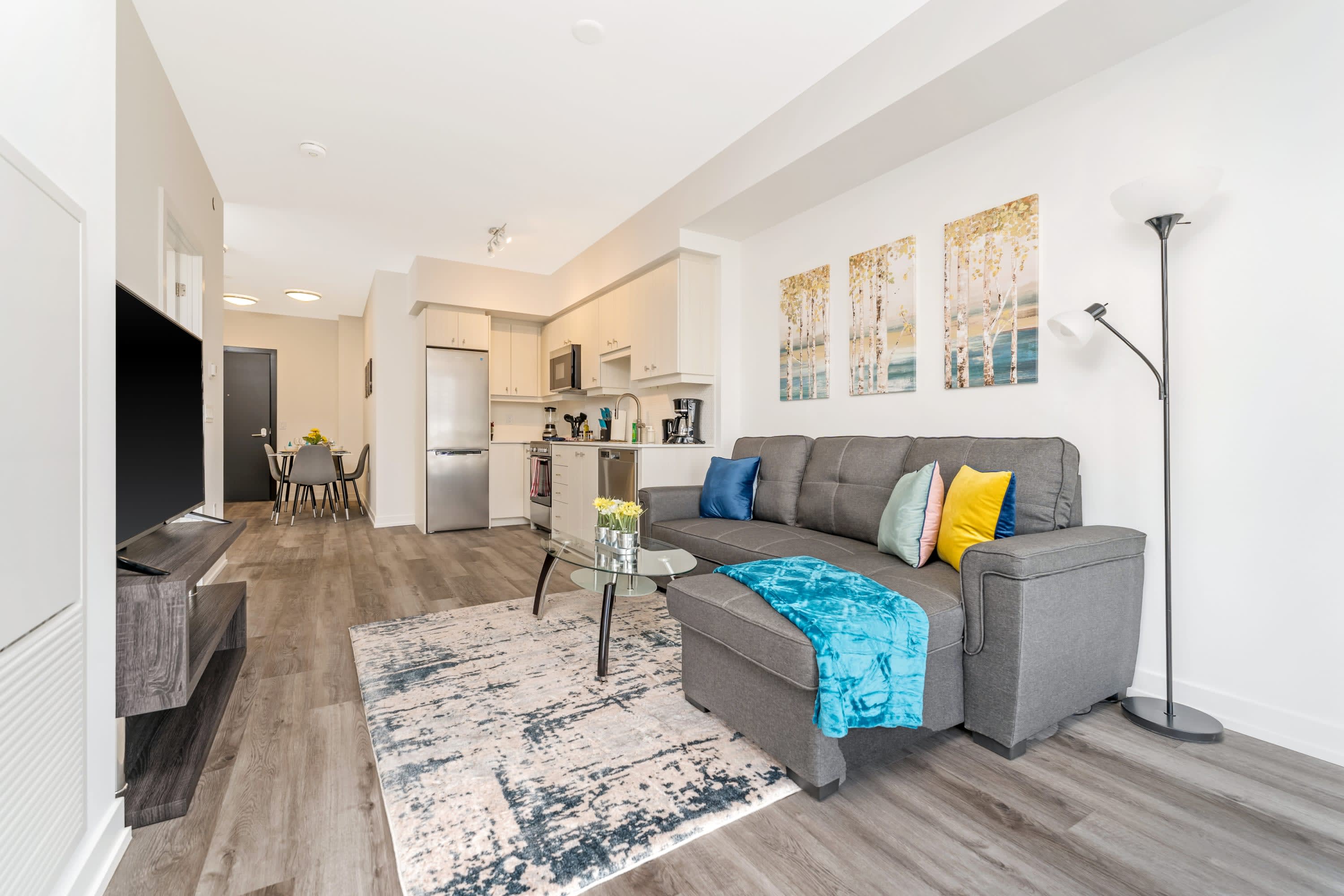 A spacious open-concept living room with a gray sectional sofa, a glass coffee table, and a large TV on a dark media console. The room features light wood-look flooring, a patterned area rug, and artwork depicting birch trees on the wall.