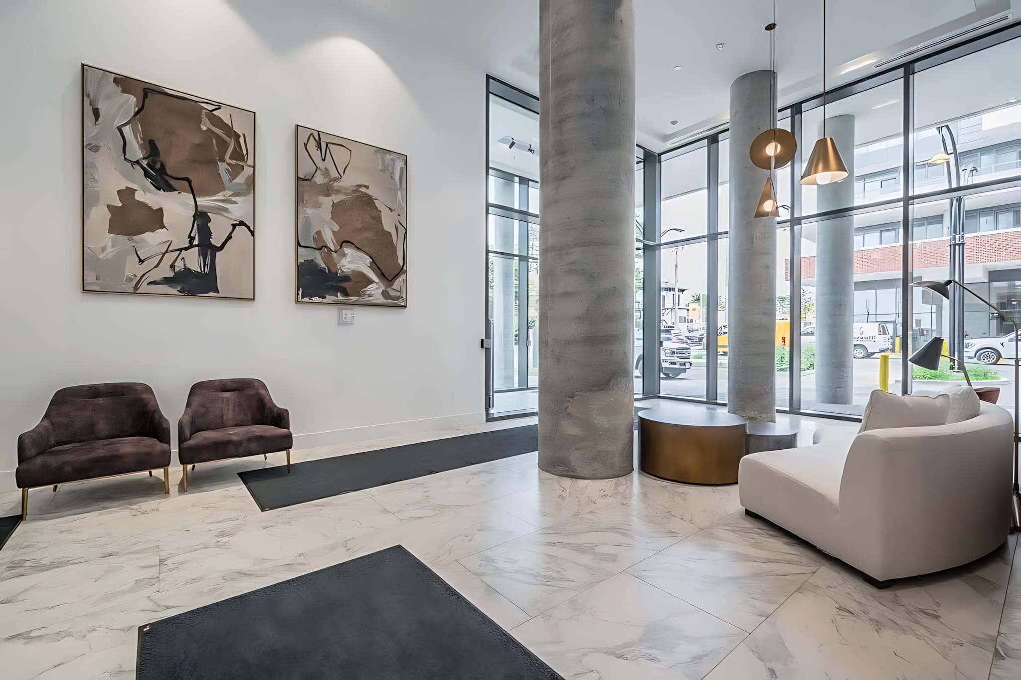 Elegant building lobby with high ceilings, large concrete pillars, and floor-to-ceiling windows. Modern artwork adorns the walls, and there are two dark armchairs and a white curved bench for seating.