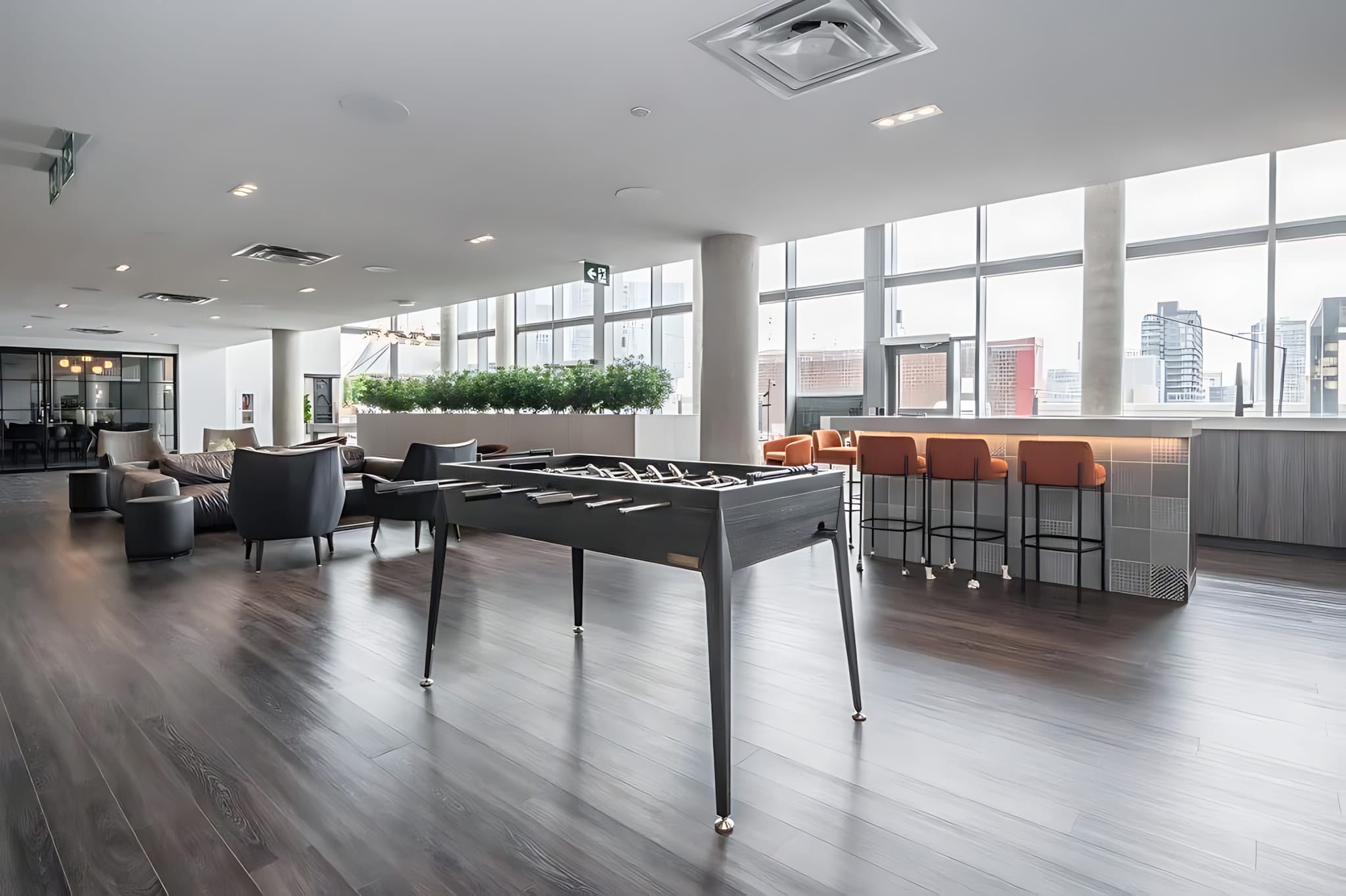A spacious and modern common lounge area featuring a sleek black foosball table as the centerpiece, surrounded by comfortable seating and a stylish bar area with orange stools. Large windows offer city views, and the room has dark wood-look flooring.