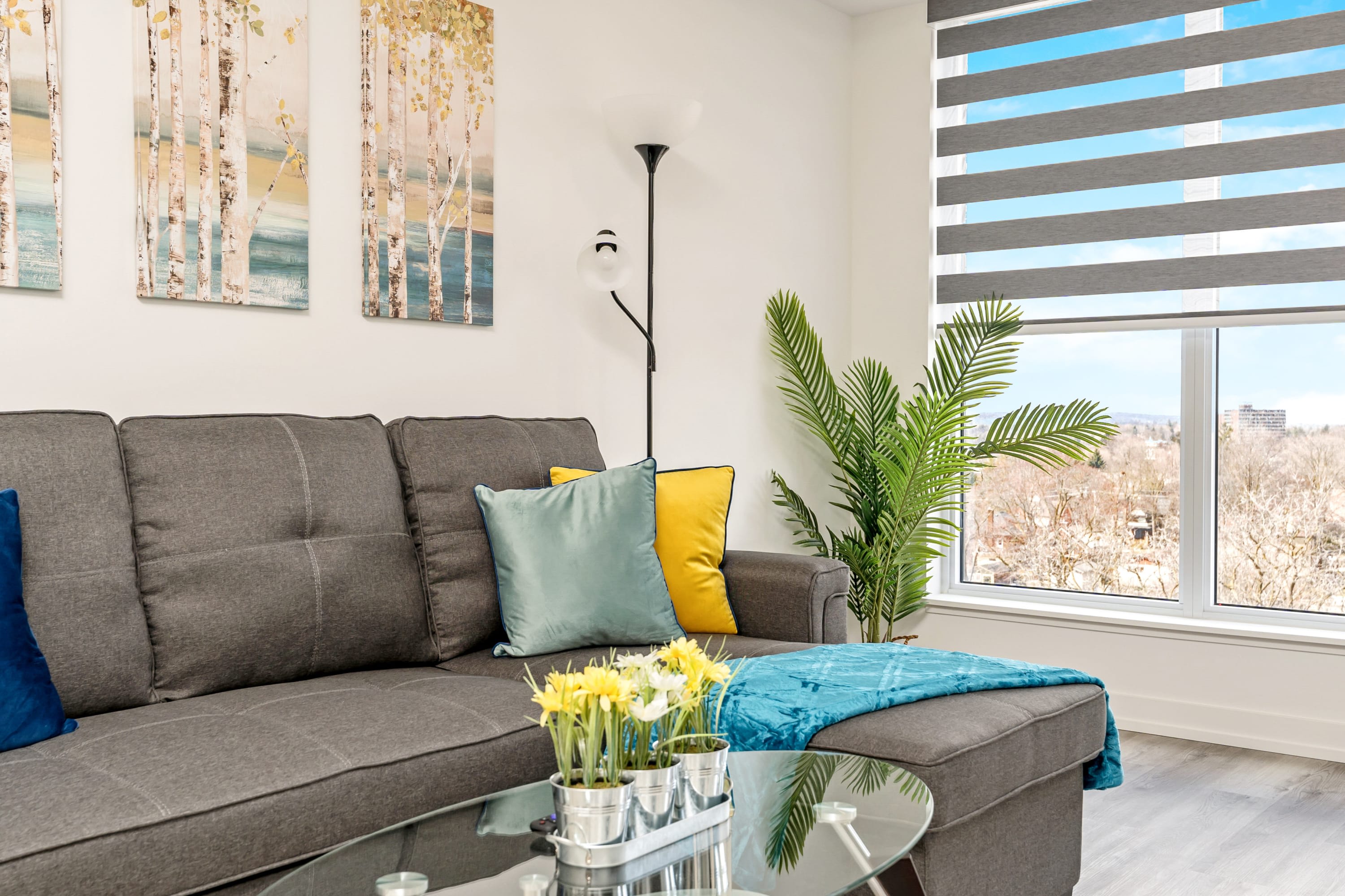 A detailed shot of the living room corner, showcasing the gray sectional sofa with light blue and yellow throw pillows, a teal blanket, and a glass coffee table with a floral centerpiece. A tall floor lamp and a large potted plant are positioned next to the sofa, with birch tree artwork on the wall.