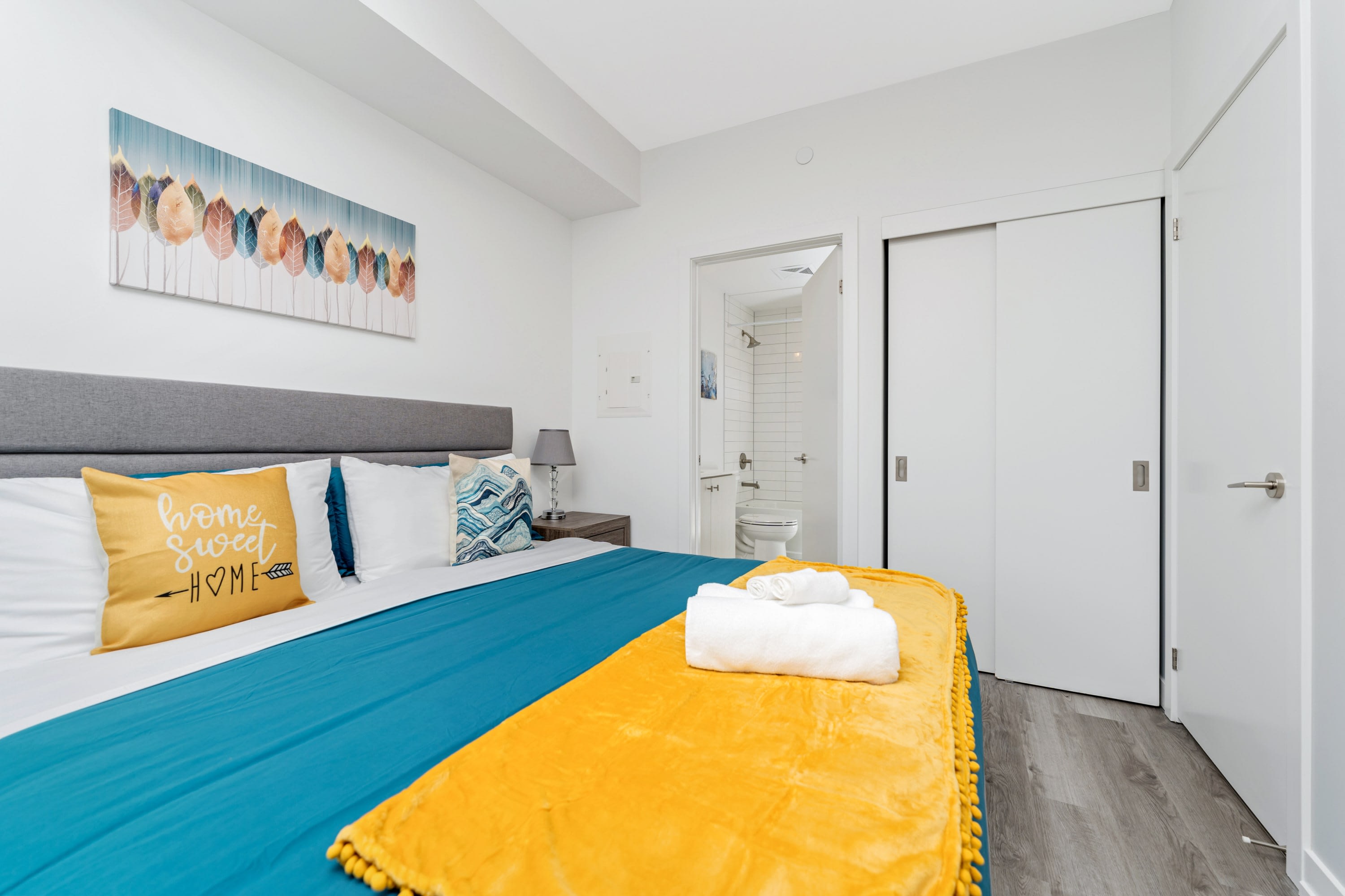 Bright bedroom with a queen-sized bed featuring a gray headboard, blue duvet, and yellow throw blanket. A colorful abstract painting hangs above the bed, and a bathroom is visible through an open doorway.