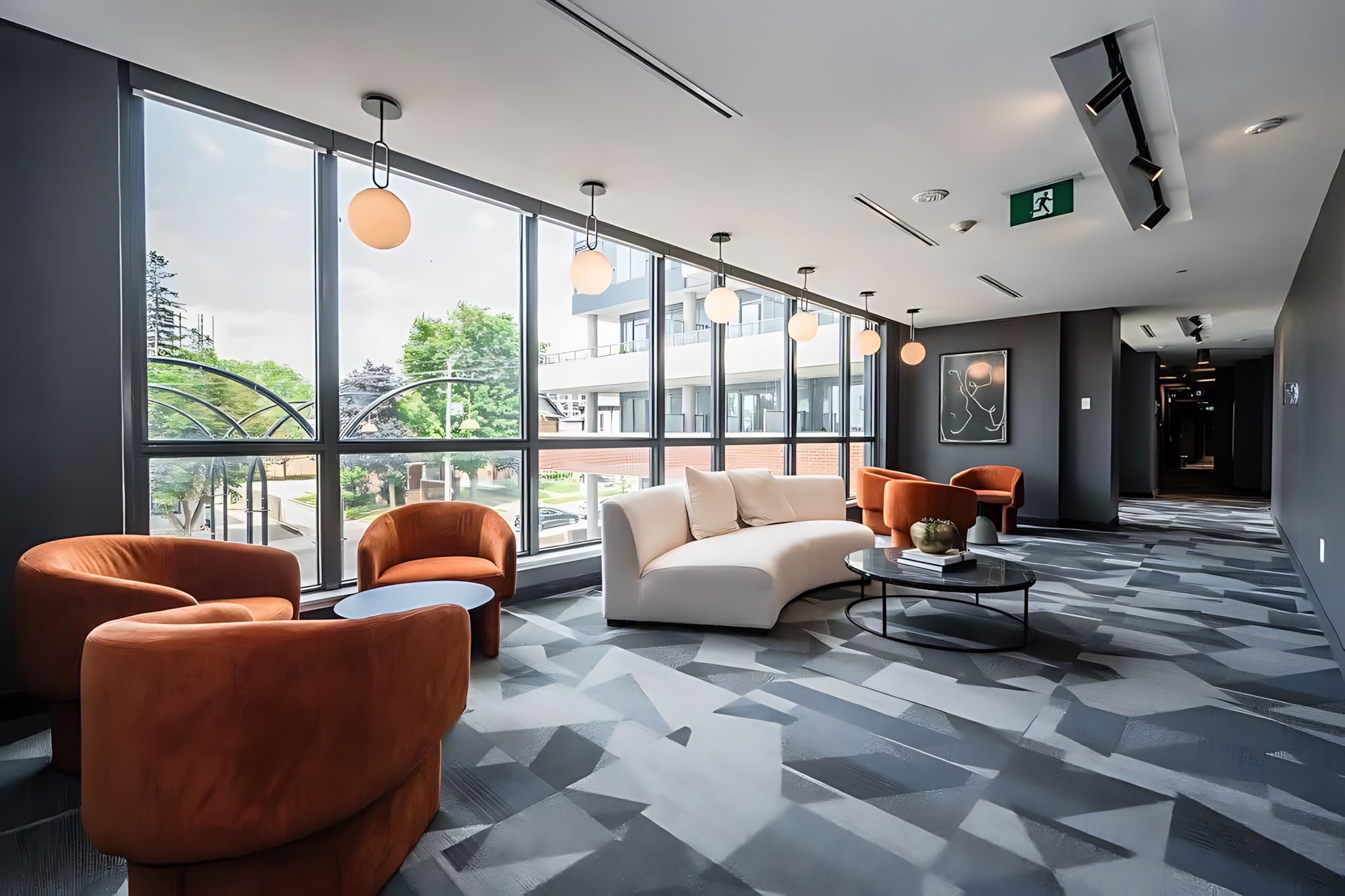 Stylish common lounge area with large windows, a curved white sofa, and several burnt orange armchairs. The floor features a striking geometric pattern in gray tones.