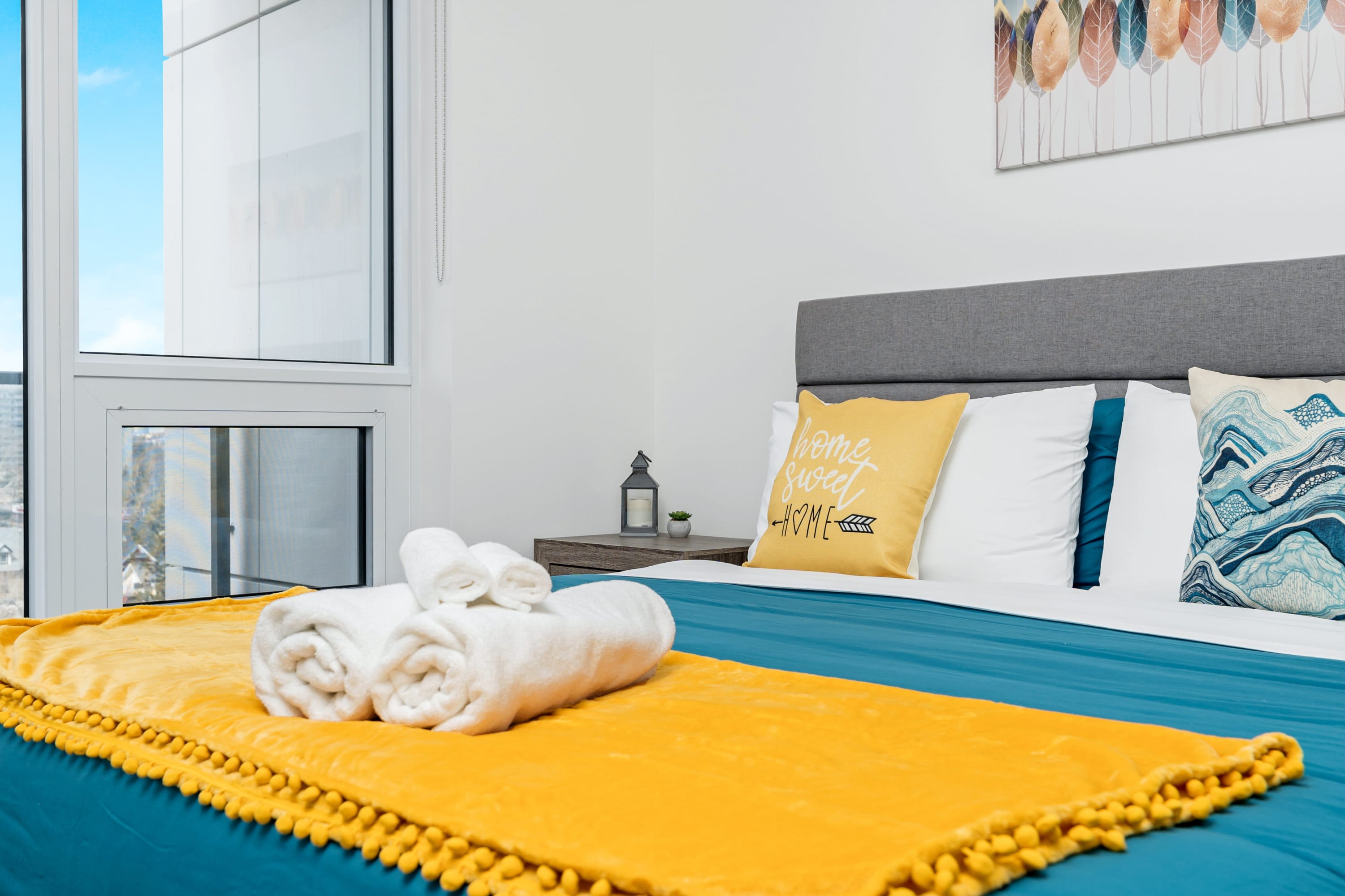 Close-up of a neatly made queen bed with a gray headboard, blue duvet, and yellow throw blanket with tassels. A "home sweet HOME" pillow and a large window with city views are visible.