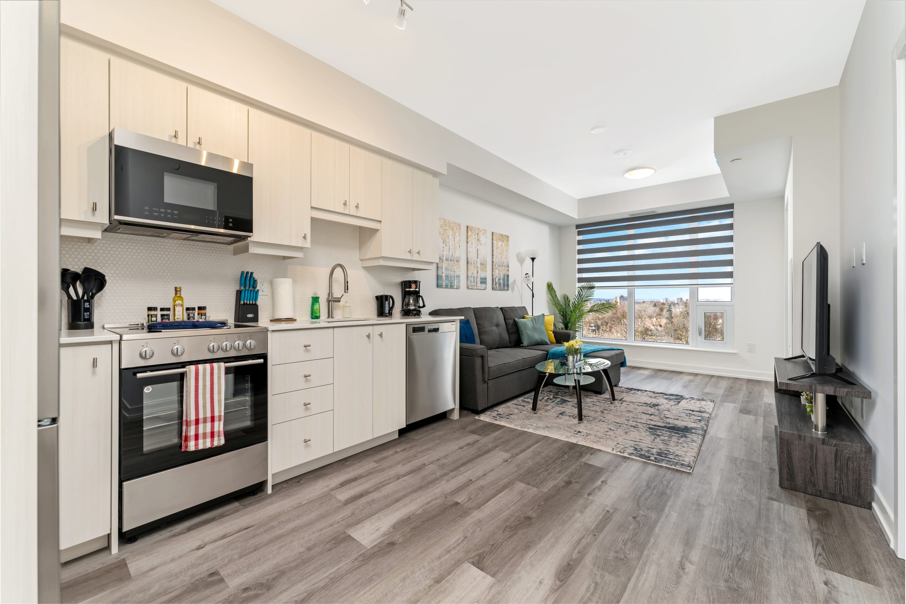 A wide shot of the open-concept living room and kitchen, featuring a gray sectional sofa with colorful pillows, a glass coffee table, and a modern kitchen with light cabinetry and stainless steel appliances. Large windows with horizontal blinds offer city views.