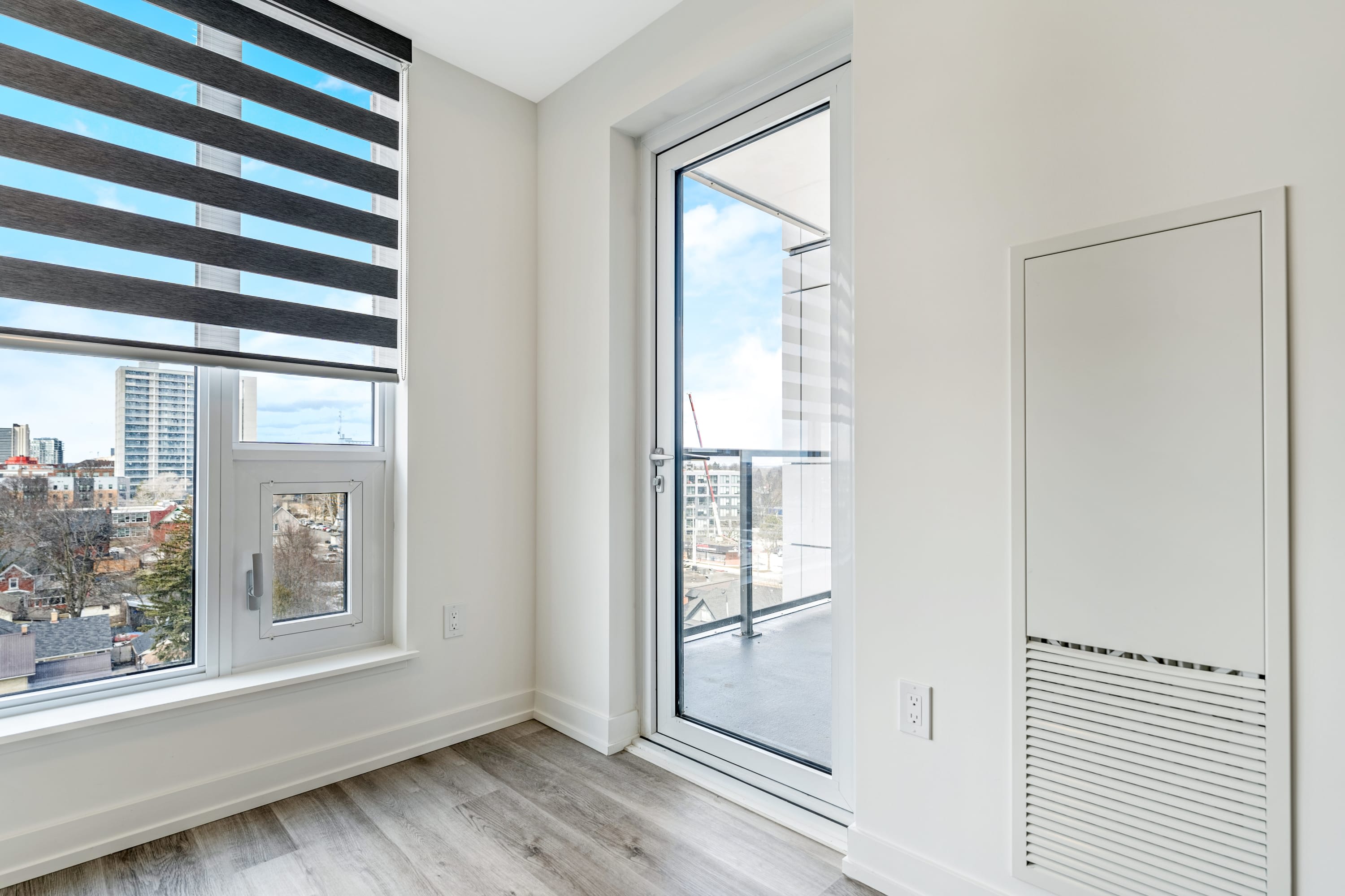 This image shows a corner of the living room with a large window featuring horizontal blinds and a glass door leading to the balcony. The room has light wood-look flooring and white walls, with a white utility panel visible on the right.