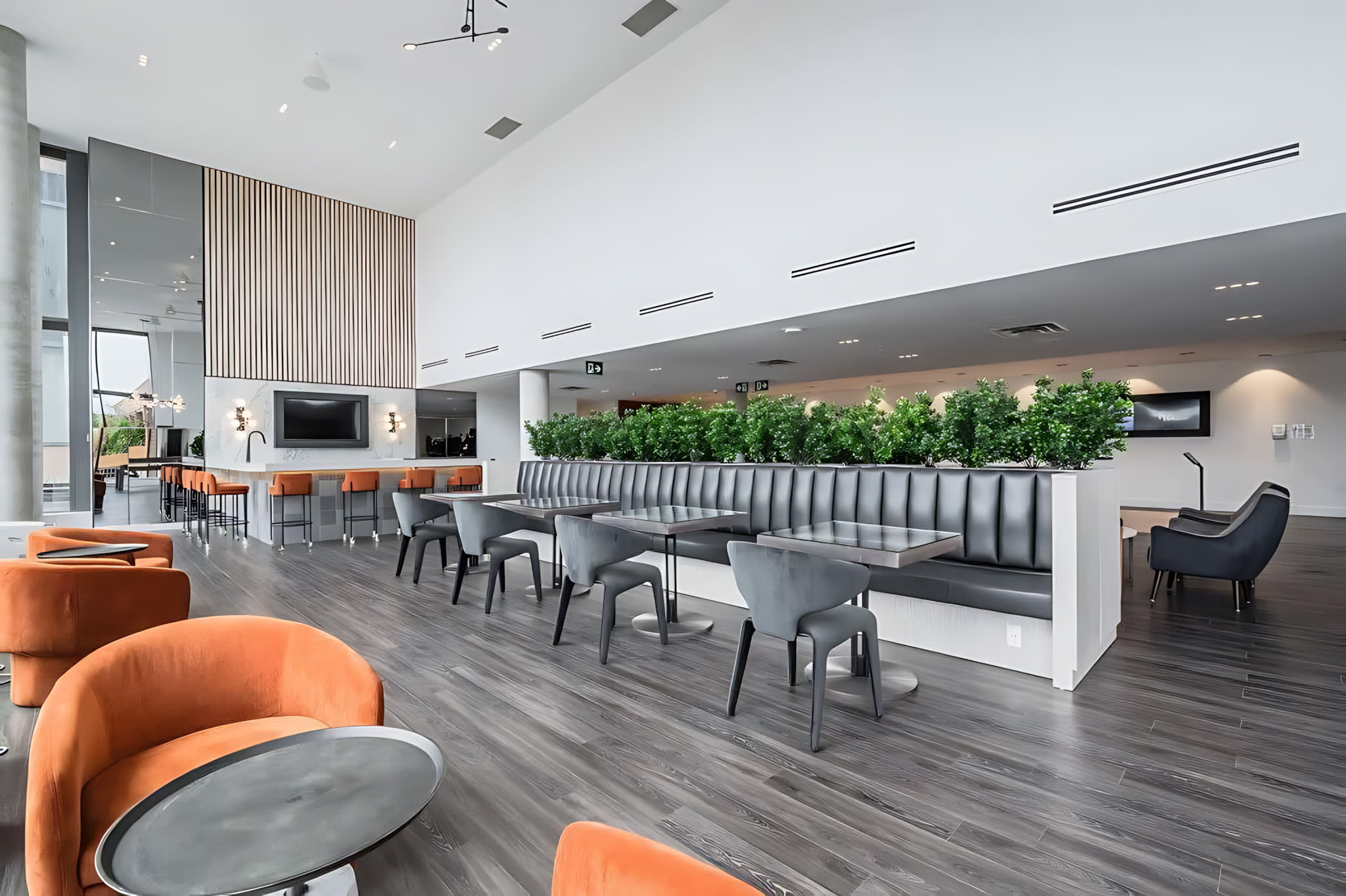 A contemporary communal lounge and dining area with varied seating options, including orange armchairs, booth seating, and small tables, alongside a bar area. Lush green plants divide the space, and large windows provide ample natural light.