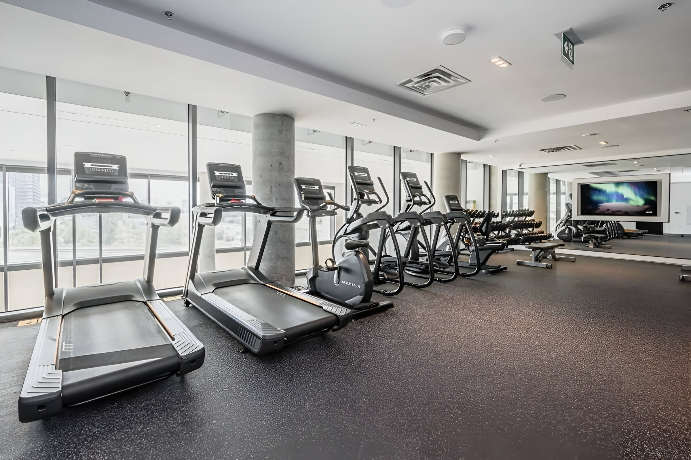 A bright, modern gym features several treadmills and elliptical machines lined up against large windows, offering city views. The floor is dark, textured rubber, and concrete pillars add an industrial touch.