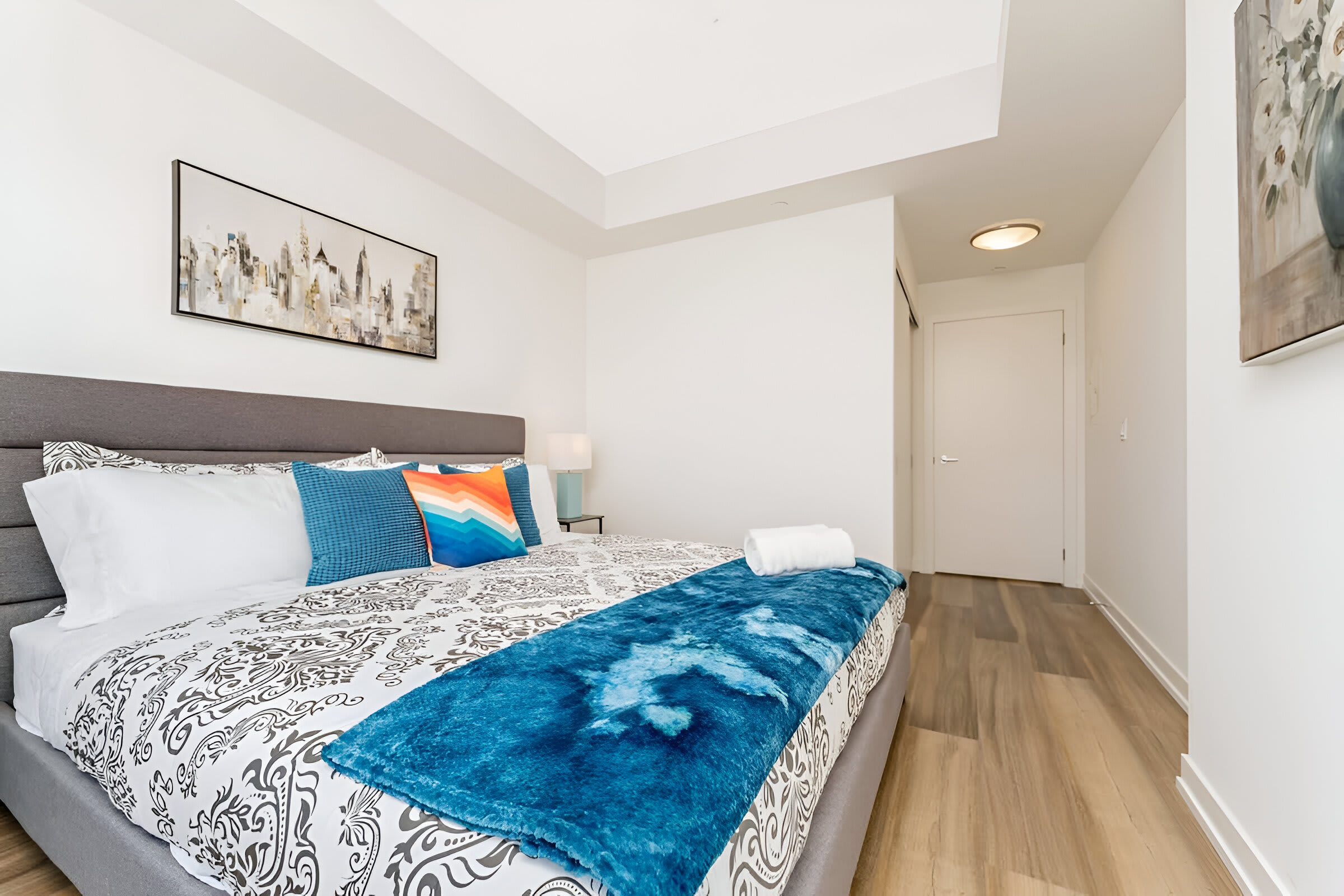 A modern bedroom showcases a gray upholstered king bed with a patterned duvet and vibrant blue and orange accent pillows, set against white walls. Light wood flooring extends throughout the room, with a glimpse of a closet and a hallway beyond.