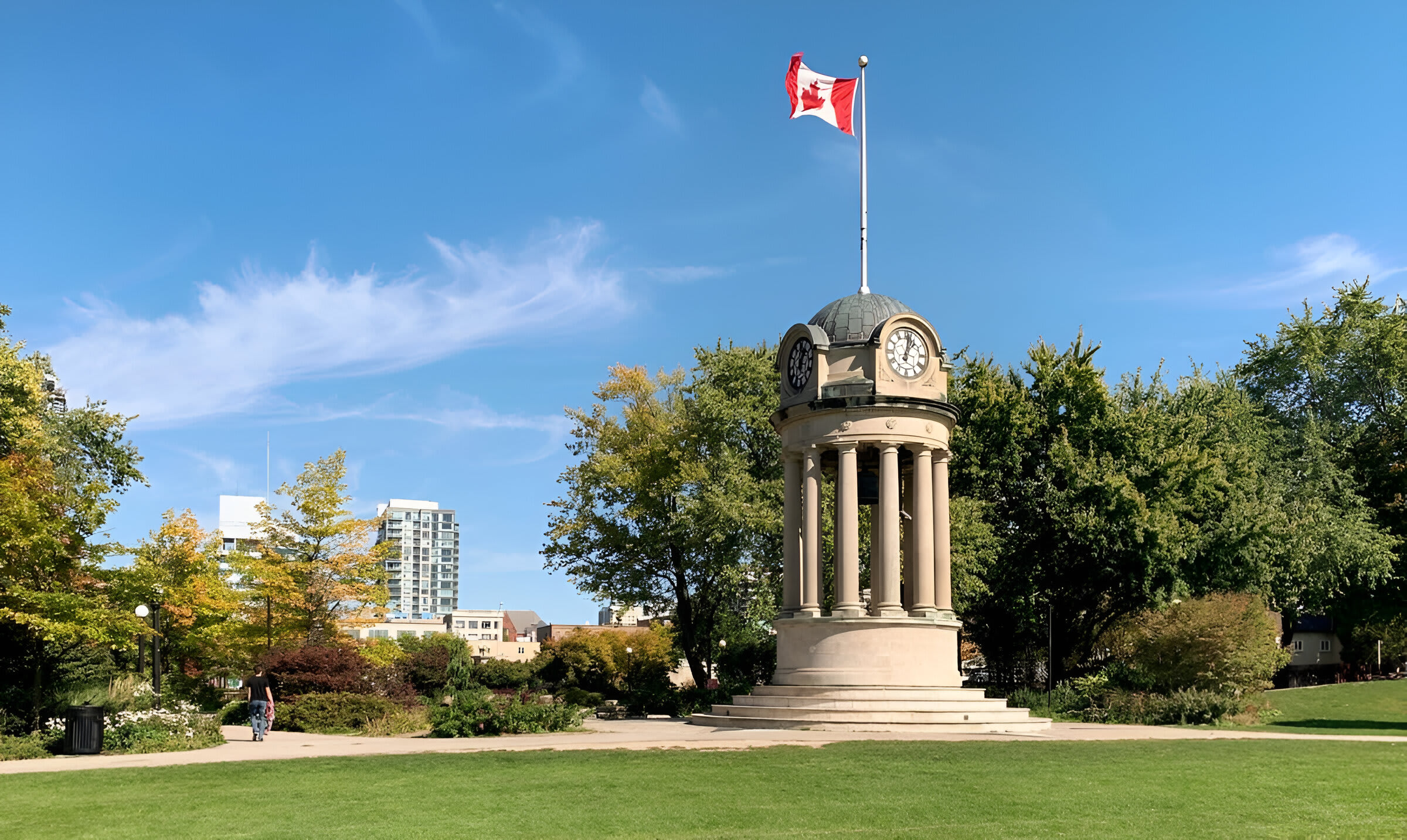 A sunny park scene features a grand stone clock tower monument topped with a Canadian flag, surrounded by lush green grass and mature trees with autumn foliage. Modern city buildings are visible in the background.