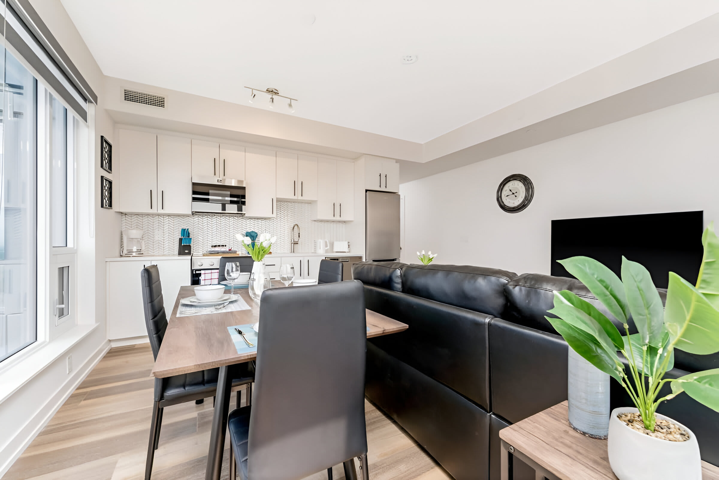 An open-concept space combines a modern kitchen with white cabinetry and stainless steel appliances, a dining area with a dark wood table, and a black leather sectional sofa. Light wood flooring unifies the bright, contemporary design.