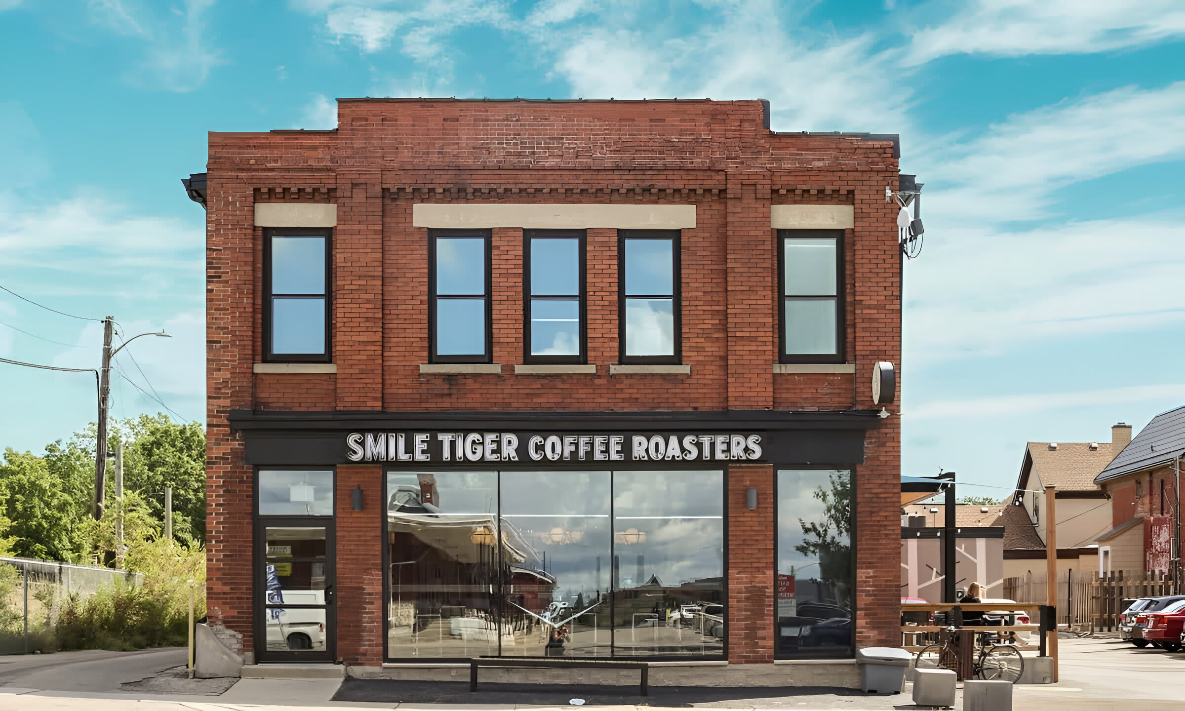 A charming brick building houses "Smile Tiger Coffee Roasters," featuring large storefront windows and a classic brick facade under a bright blue sky. A small bench and bicycle are visible outside.