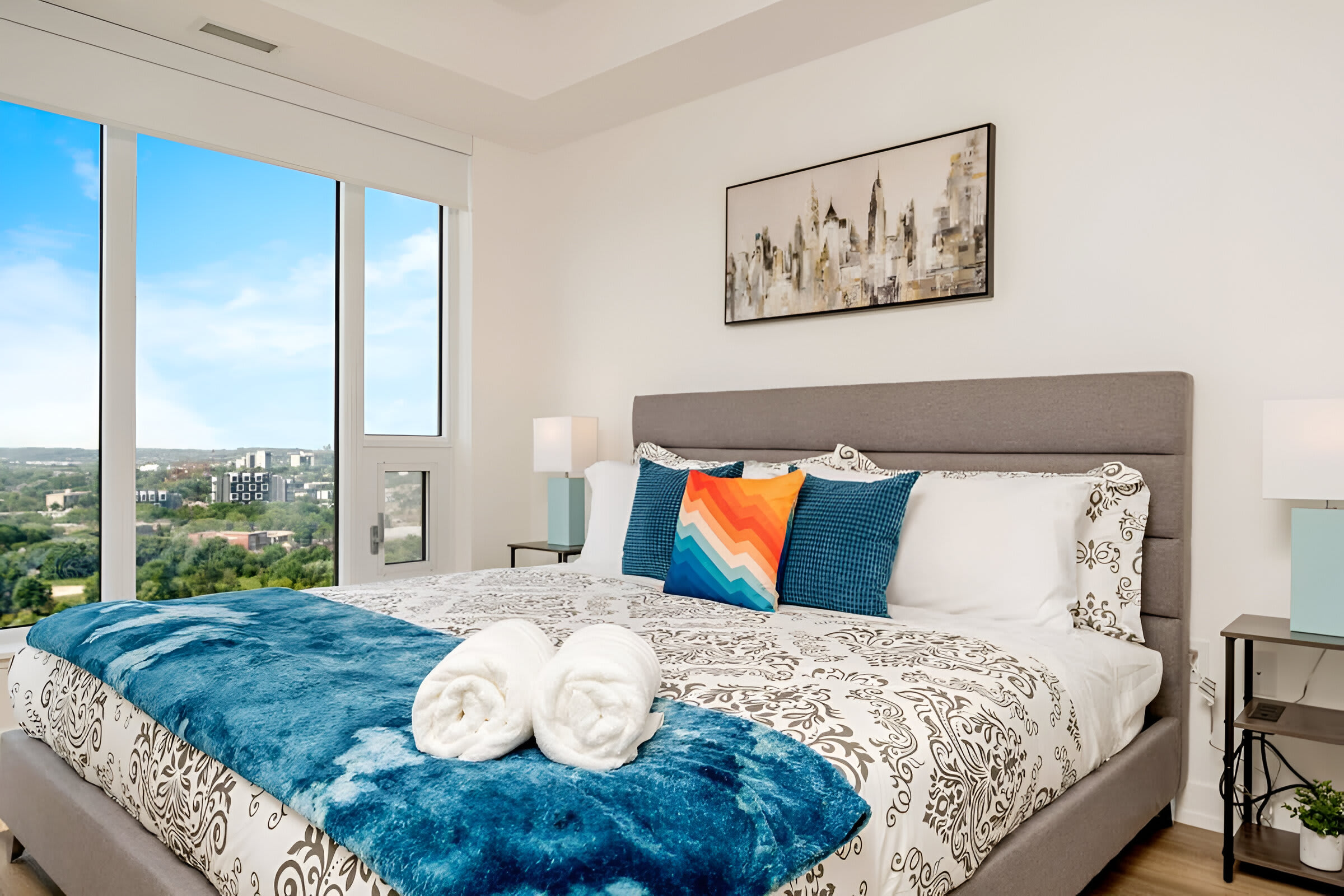 The bright bedroom features a gray upholstered king-size bed with a patterned duvet, a blue throw blanket, and colorful accent pillows. Floor-to-ceiling windows offer expansive city views, complemented by light wood flooring and a modern abstract art piece above the bed.