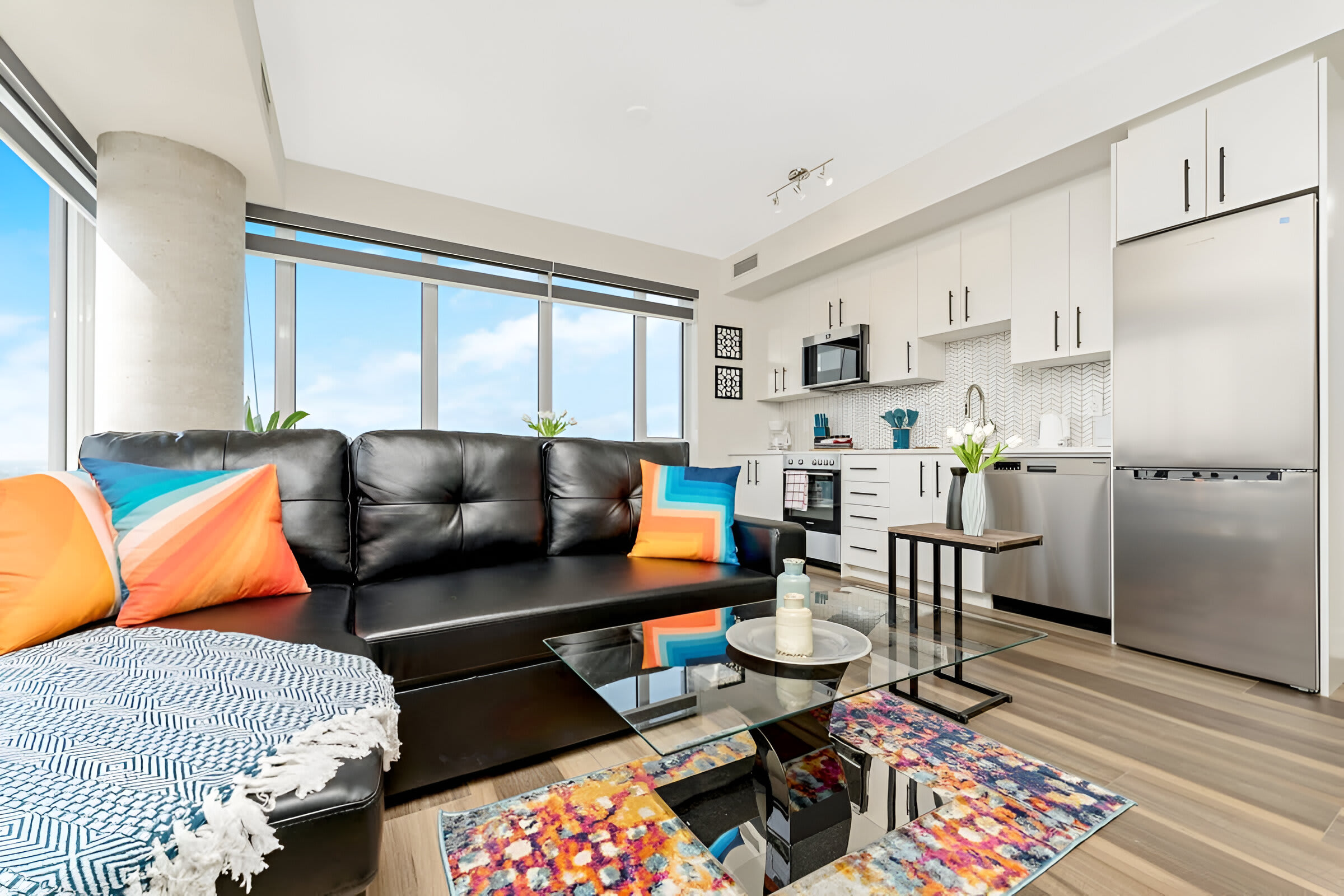 Open-concept living space combining a modern kitchen with white cabinets and stainless steel appliances, and a living area with a black leather sectional sofa and a glass coffee table, all with city views.