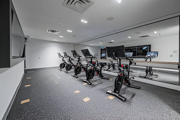 A dedicated spin studio within the gym features several stationary bikes arranged in rows, facing a large mirrored wall. The room has dark, textured flooring and recessed lighting.