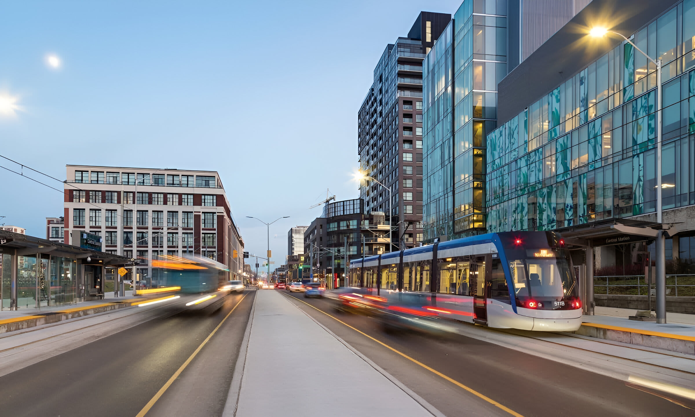 A vibrant city street scene at dusk shows a modern tram moving along tracks, flanked by contemporary buildings with large glass windows. Streetlights illuminate the busy urban environment.