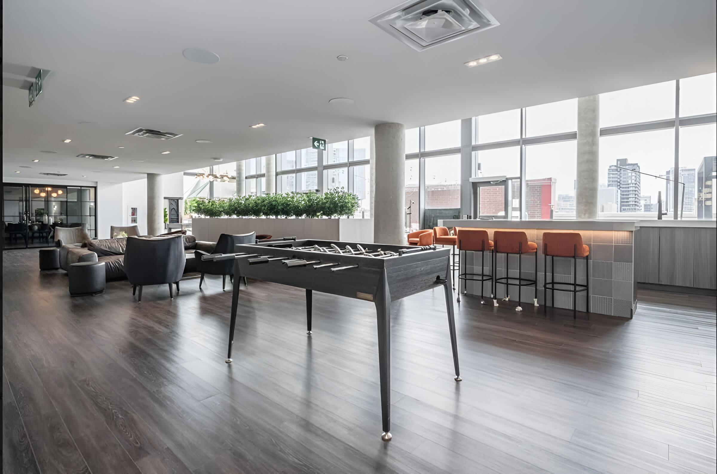 A spacious common area features a foosball table, various seating arrangements including modern armchairs and a long bench with tables, and large windows offering city views. The room has dark wood flooring and a contemporary design.