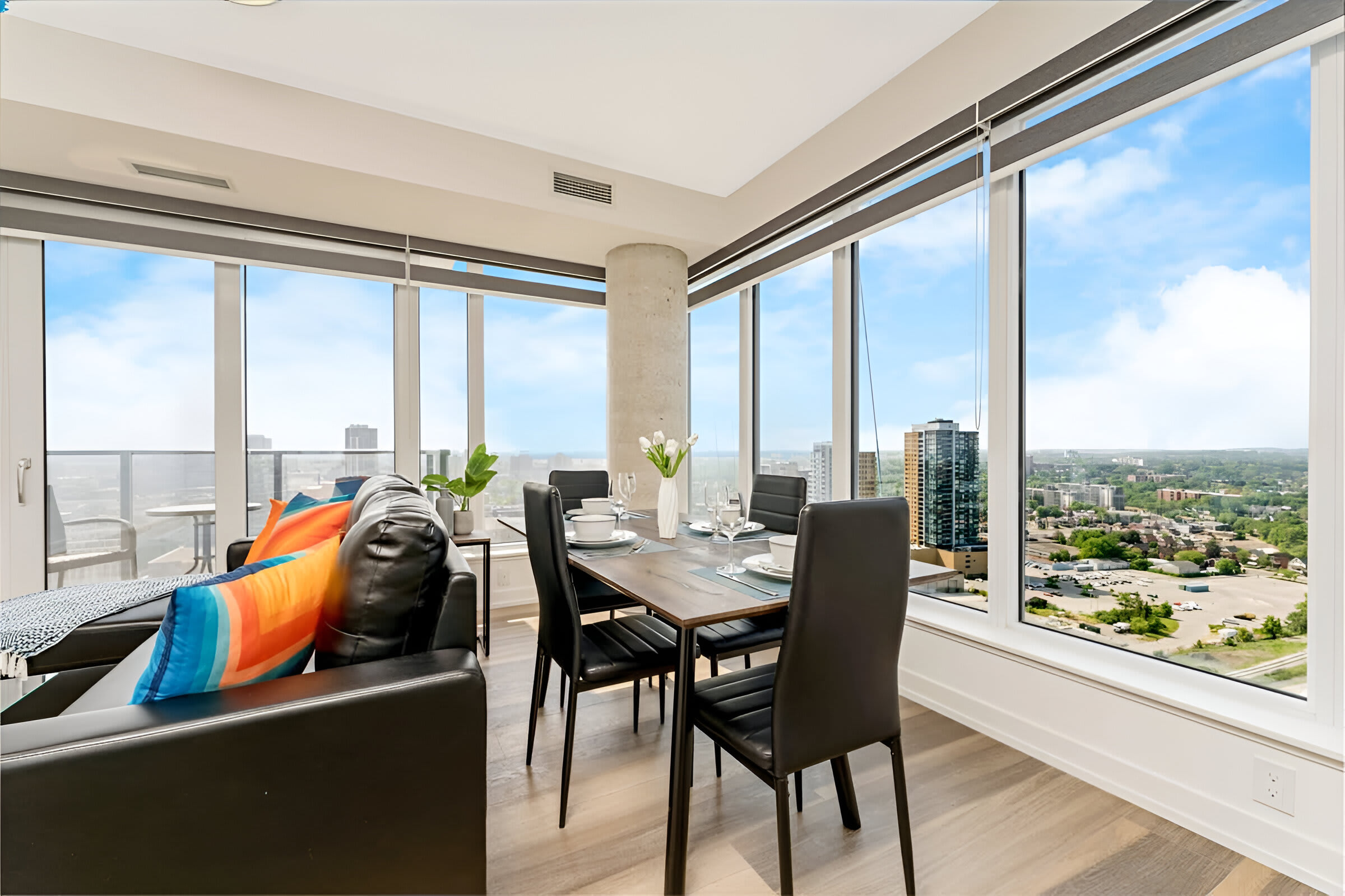A modern open-concept living and dining area features a black leather sofa with colorful throw pillows, a dark wood dining table with black chairs, and floor-to-ceiling windows offering panoramic city views. Light wood flooring and a concrete pillar add to the contemporary aesthetic.