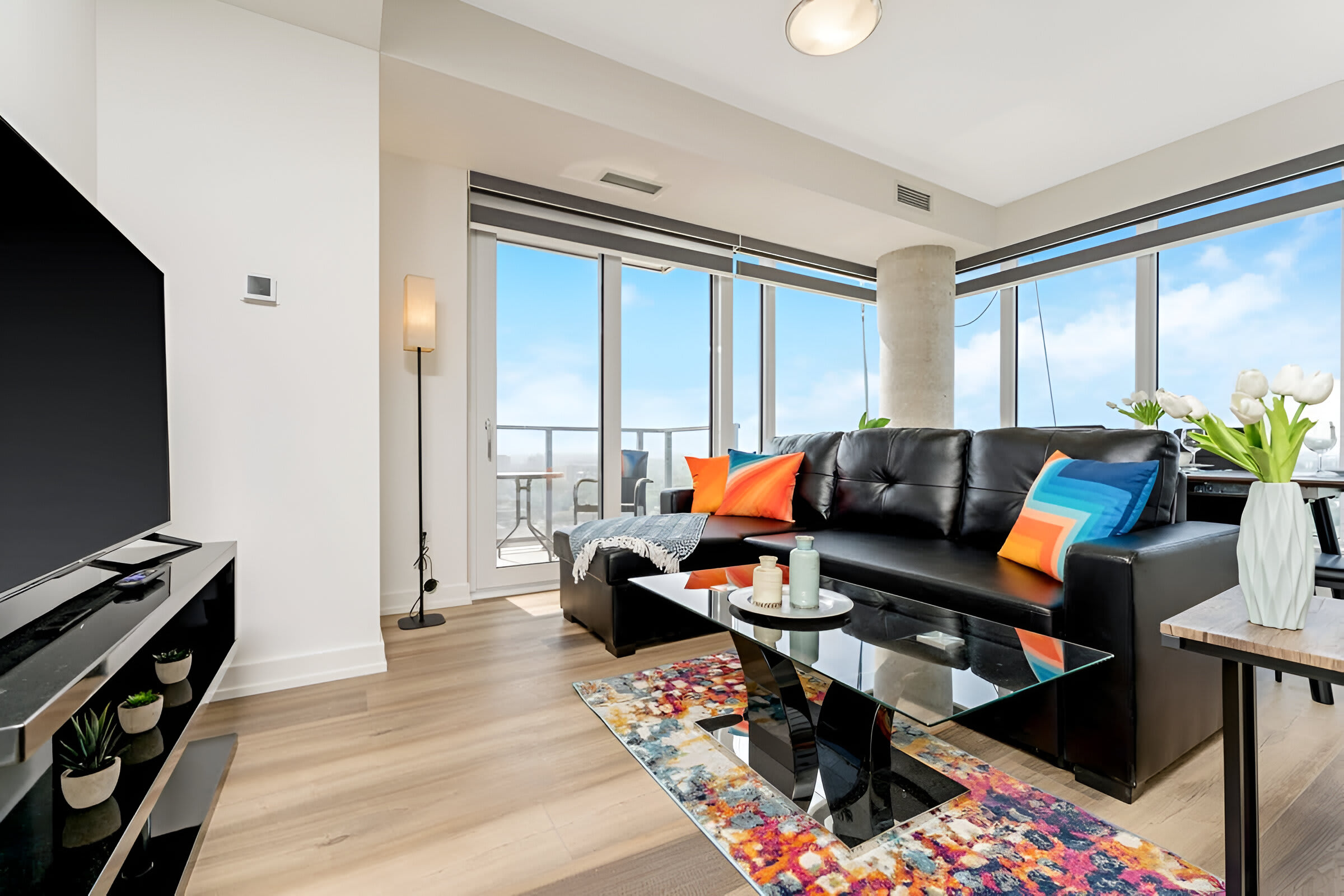 Contemporary living room featuring a large flat-screen TV on a black console, a black leather sectional sofa with colorful pillows, and a glass coffee table, with large windows providing city views.