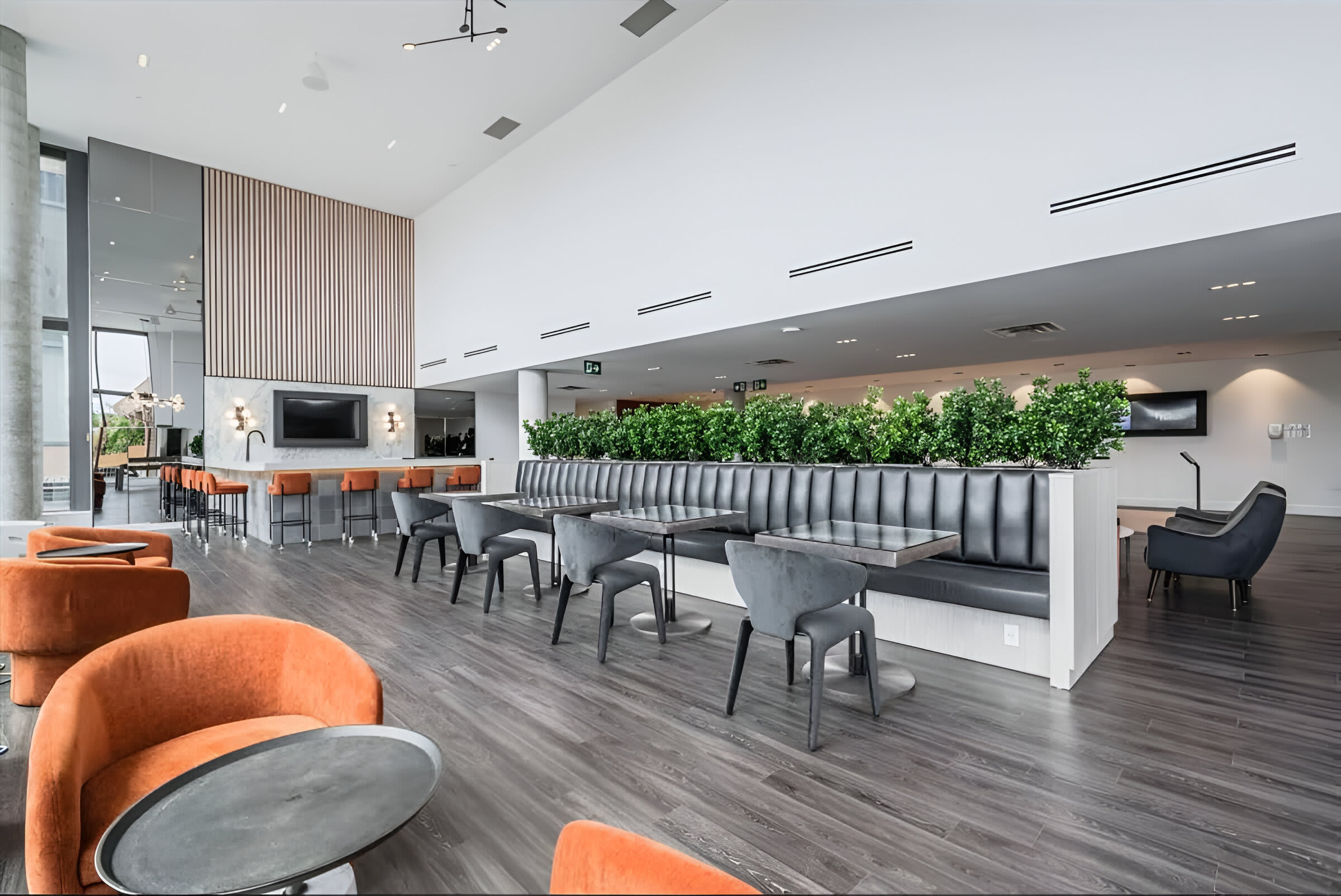 A modern common lounge area features a long banquette with individual tables and chairs, orange accent chairs, and a bar area with stools. A slatted wood accent wall and large windows create a bright and inviting space.