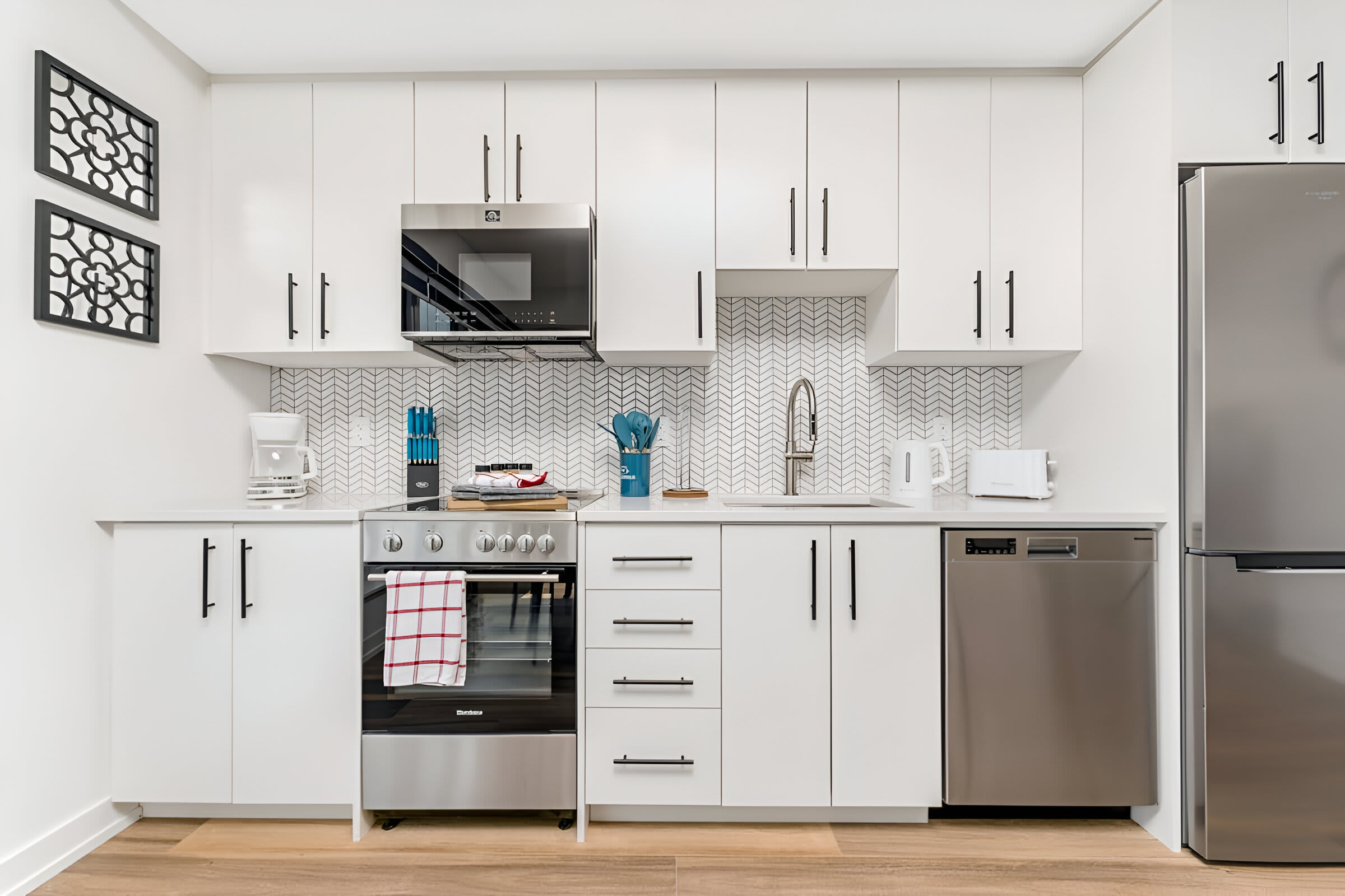 A sleek, modern kitchen features white shaker-style cabinets with black hardware, a white herringbone tile backsplash, and stainless steel appliances including a range, microwave, dishwasher, and refrigerator. A white countertop and light wood flooring complete the clean aesthetic.
