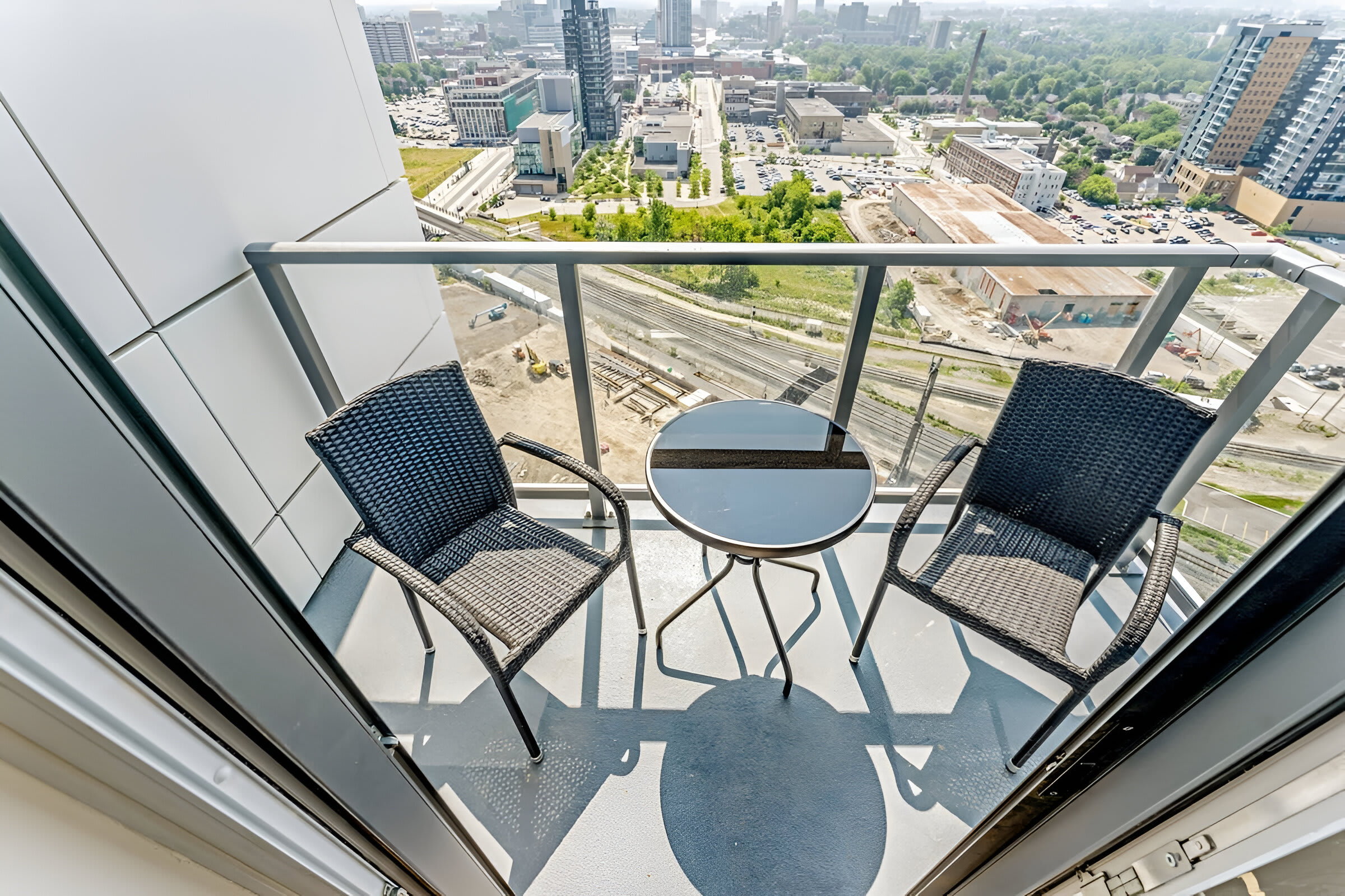 High-angle view from a balcony with a small round glass table and two chairs, showcasing a detailed urban landscape with roads, buildings, and train tracks below.