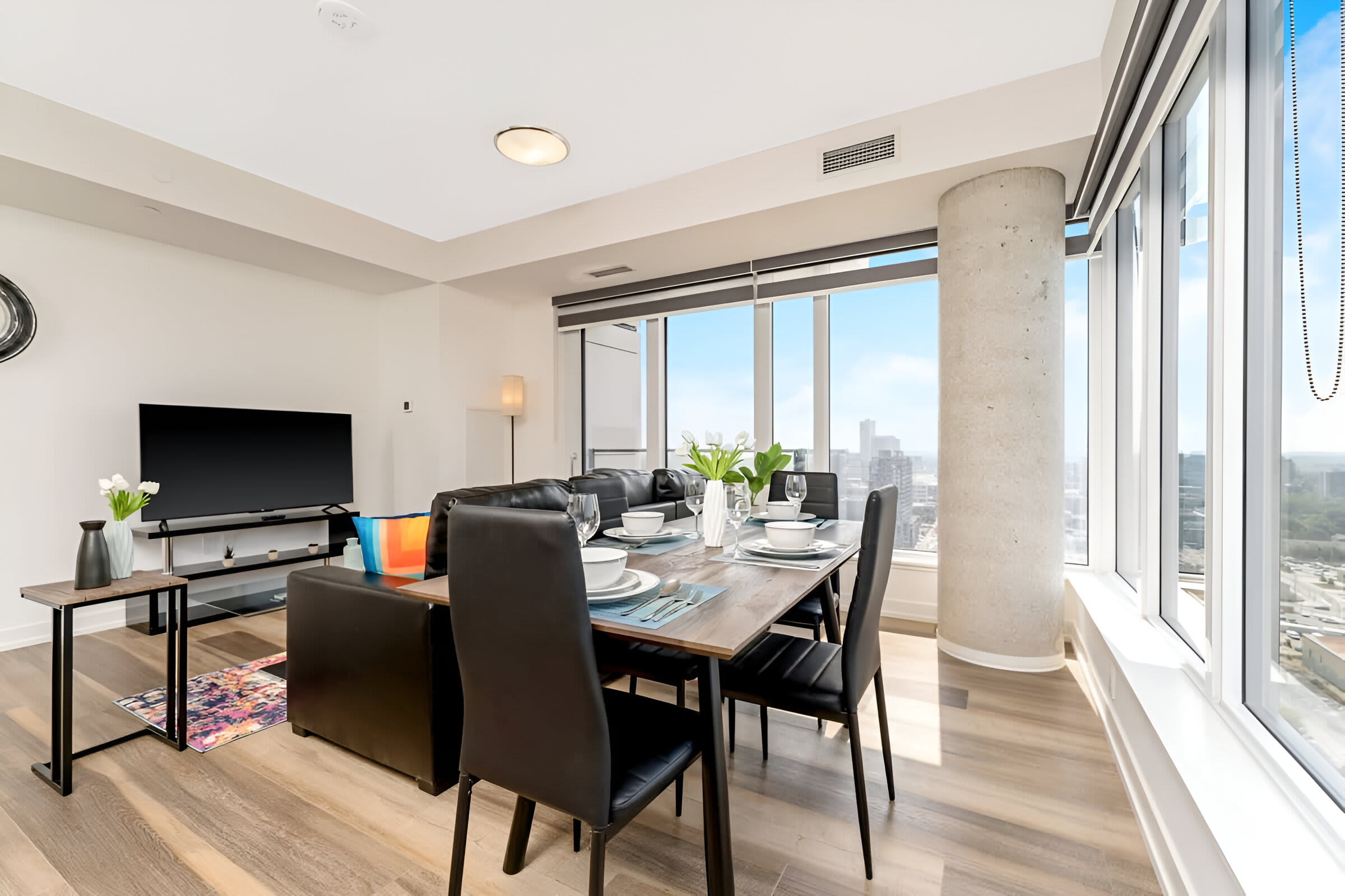 Spacious open-plan living and dining area with a black leather sofa, a dark wood dining table set for four, and a large flat-screen TV, all illuminated by natural light from expansive city-view windows.