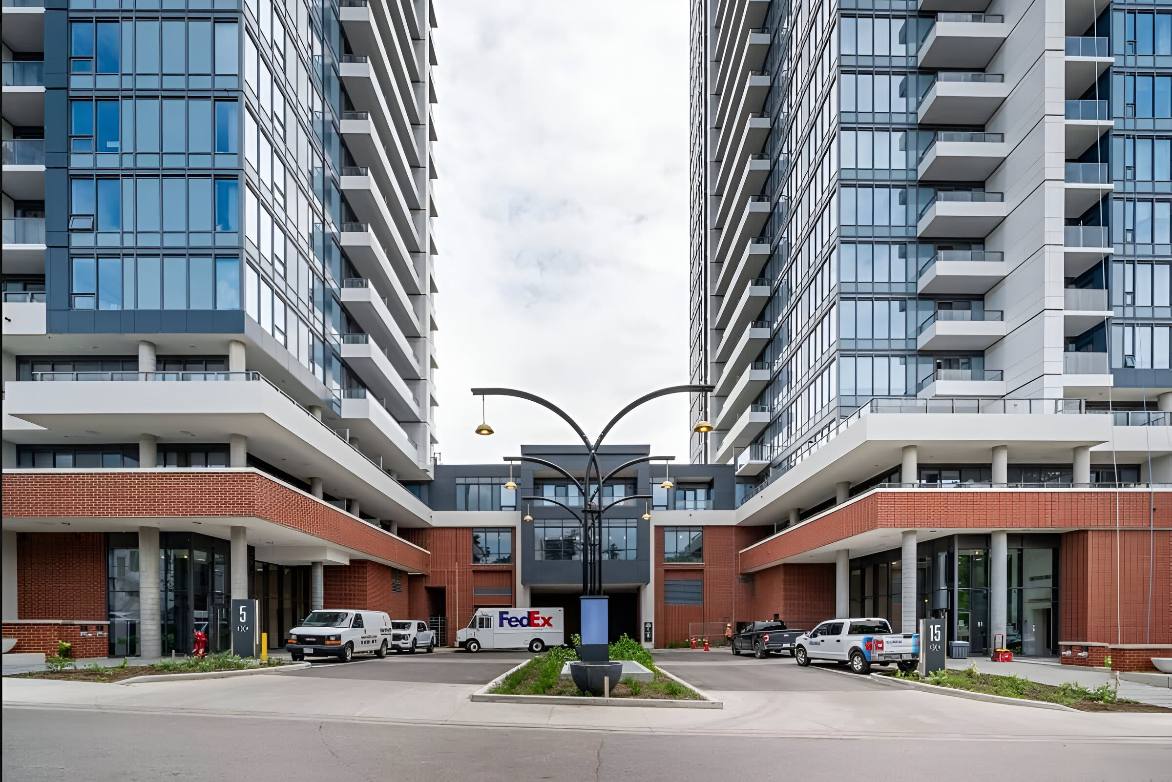The exterior view shows two modern high-rise apartment buildings with glass facades and balconies, connected by a lower central structure. A paved driveway leads to the building entrances, with delivery vehicles parked in front.
