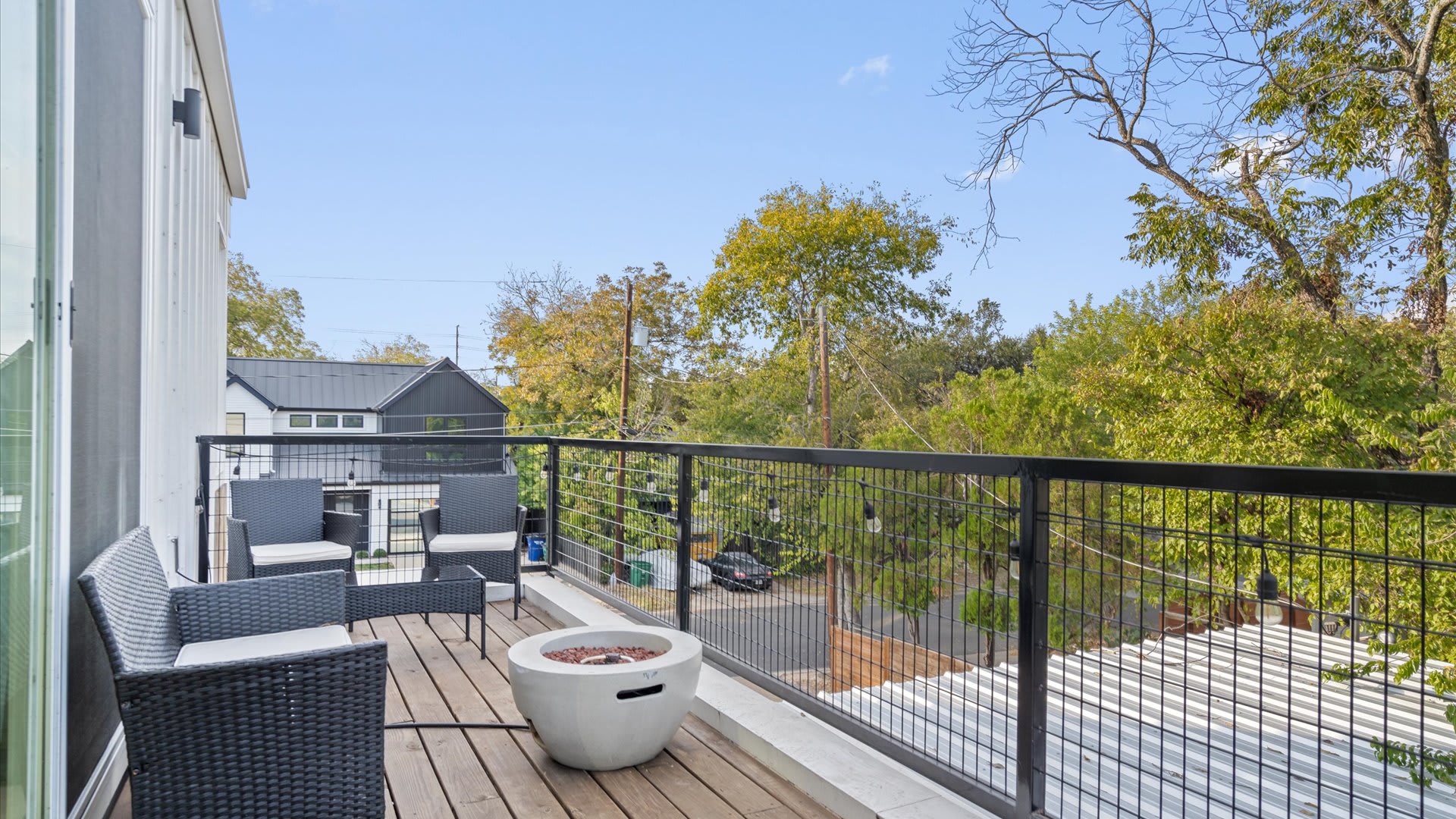 A spacious outdoor balcony featuring a black metal railing, a concrete fire pit, and dark wicker outdoor seating with white cushions, overlooking a tree-lined neighborhood.