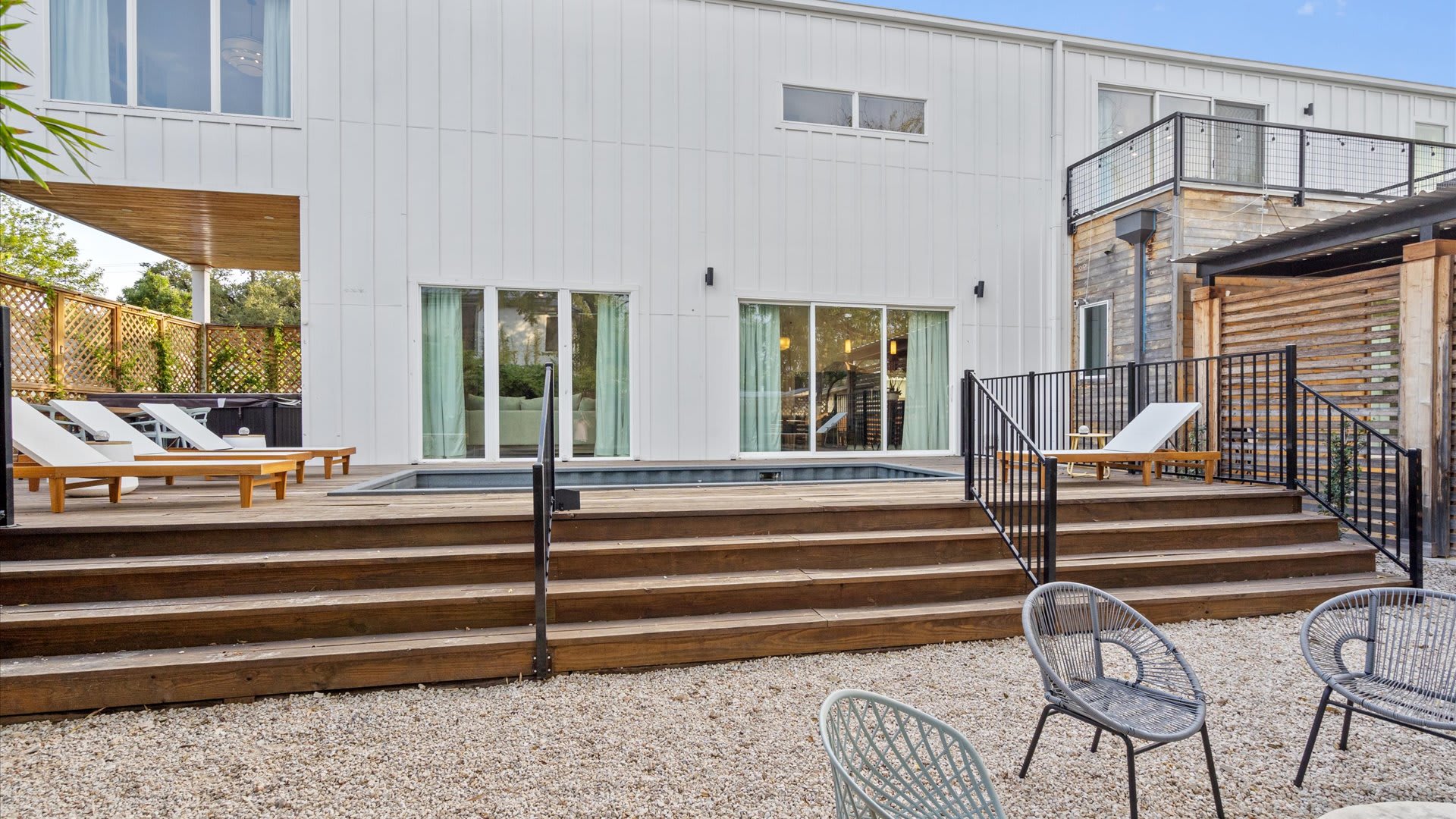 The modern white exterior of the house with large windows and glass doors, overlooking a multi-level wooden deck with a small plunge pool, lounge chairs, and a gravel seating area.
