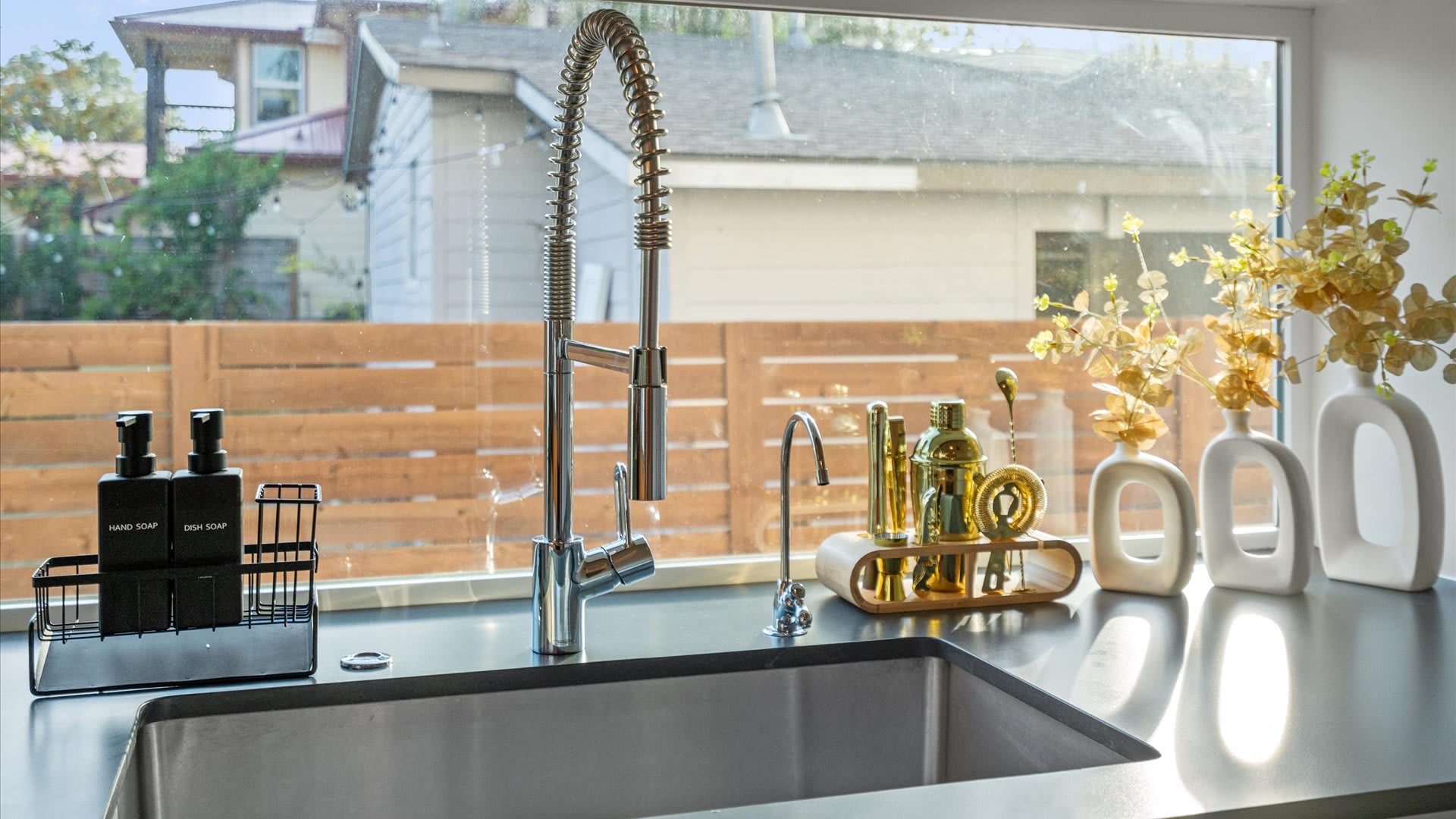 Close-up of a modern kitchen sink area with a large stainless steel basin, a flexible pull-down faucet, and a smaller filtered water tap. The dark countertop is accessorized with soap dispensers and decorative vases, overlooking a window view.