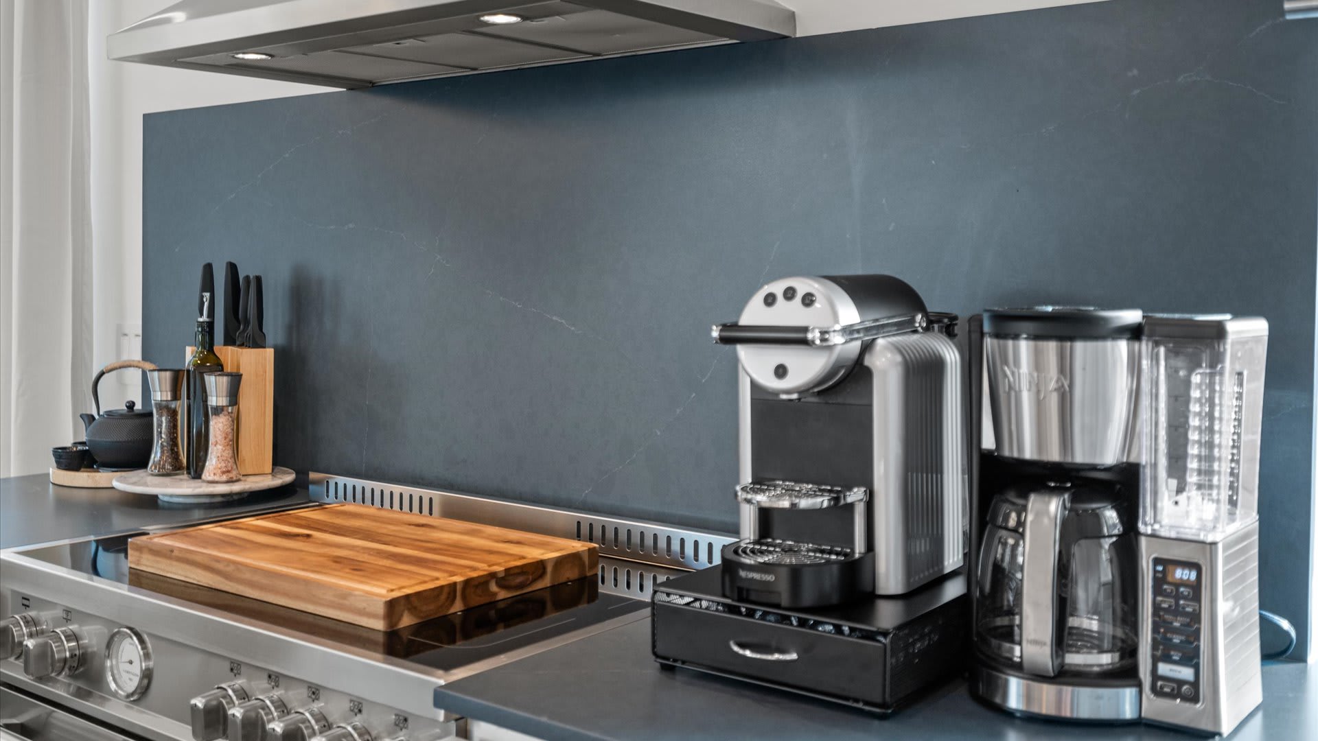 Detailed view of a modern kitchen counter featuring a stainless steel range with a wooden cutting board, flanked by a Nespresso machine and a Ninja coffee maker. A dark gray backsplash provides a sleek backdrop.
