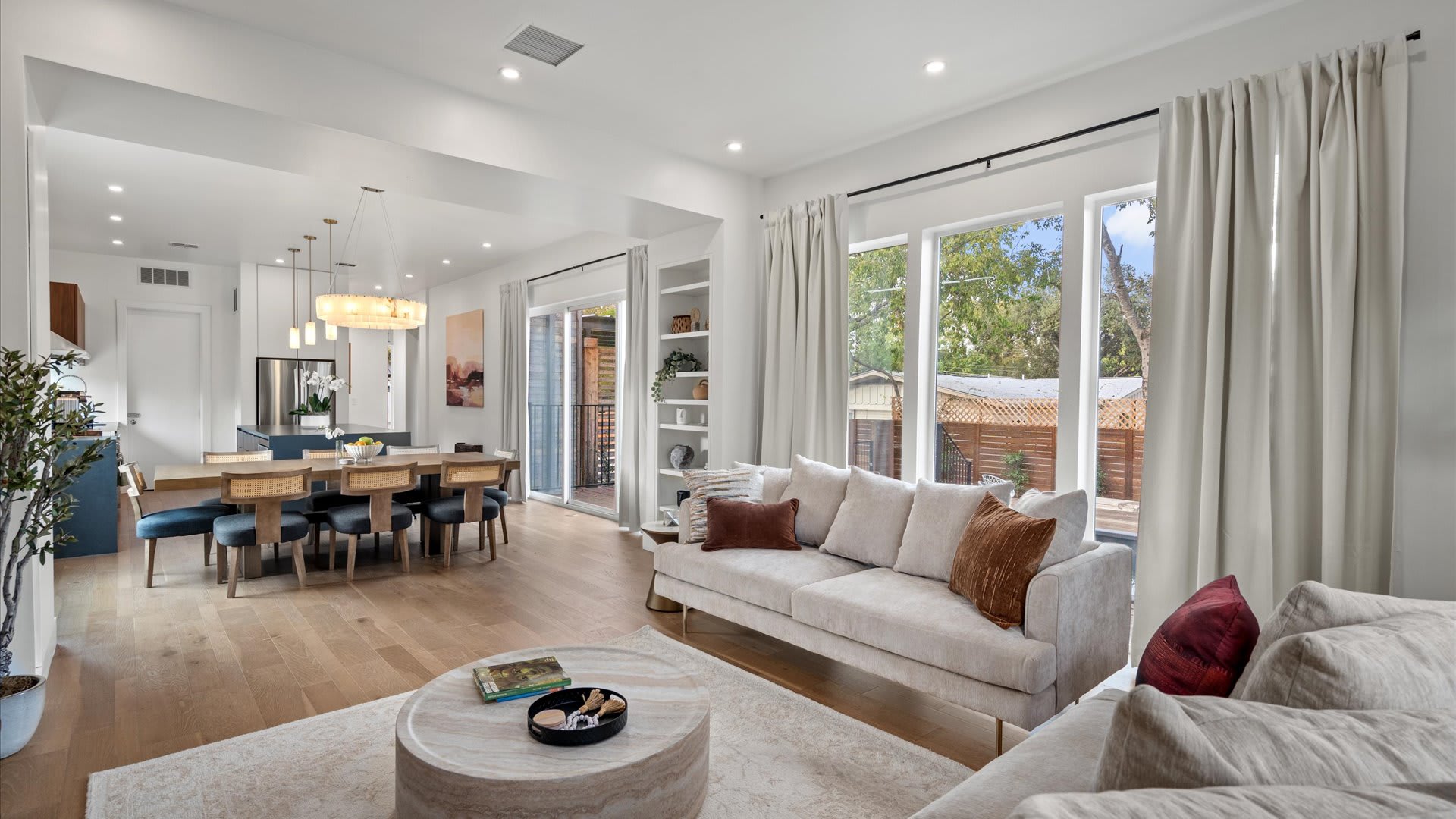 Spacious open-concept living and dining area with light hardwood floors and recessed lighting. The living room features two comfortable sofas and built-in shelves, while the dining area has a long wooden table and chairs, with a sliding glass door leading to an outdoor space.