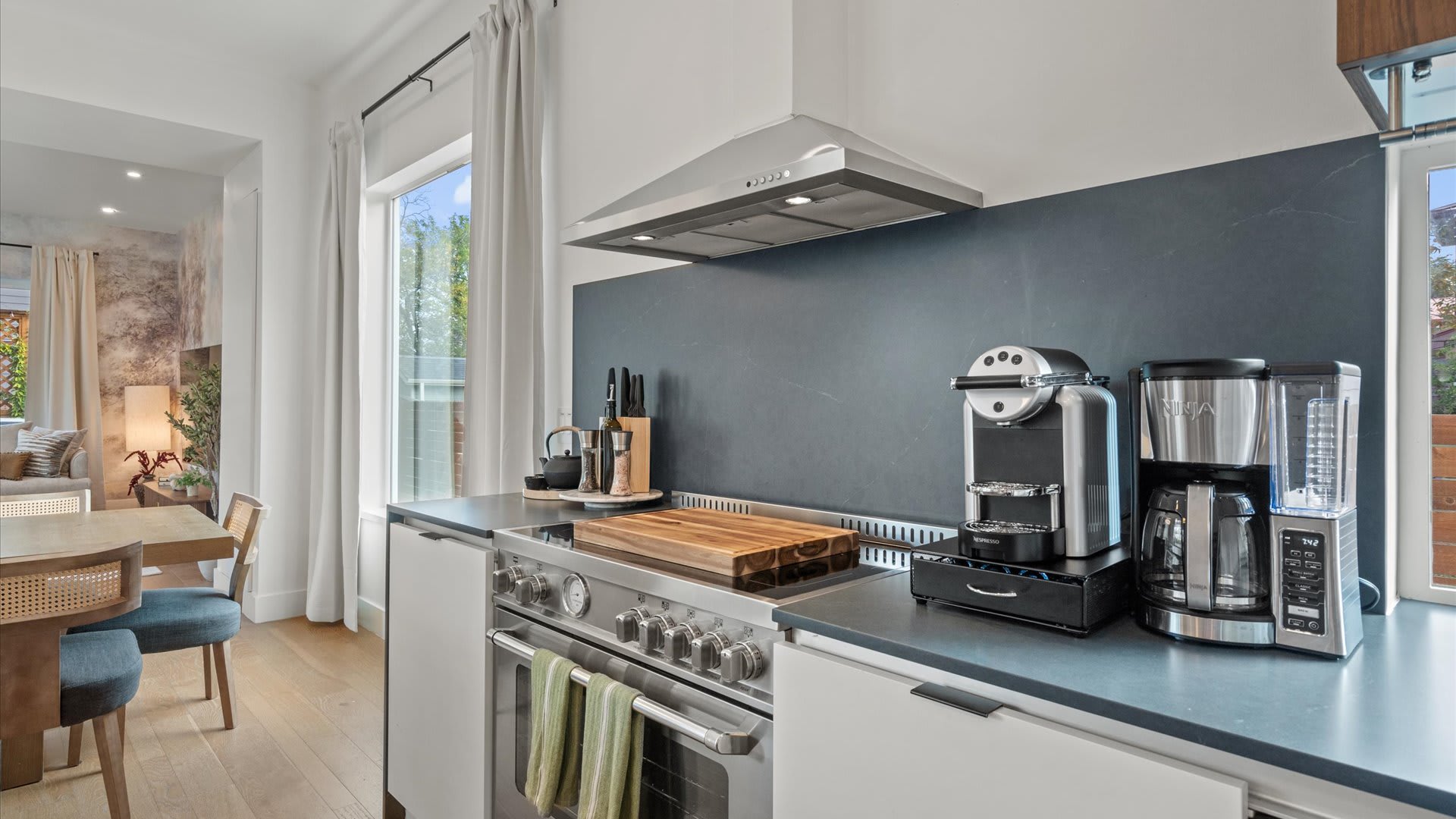 Modern kitchen featuring stainless steel appliances, a sleek dark gray backsplash, and light wood flooring. The counter holds various coffee makers and a wooden cutting board, with a range hood overhead.