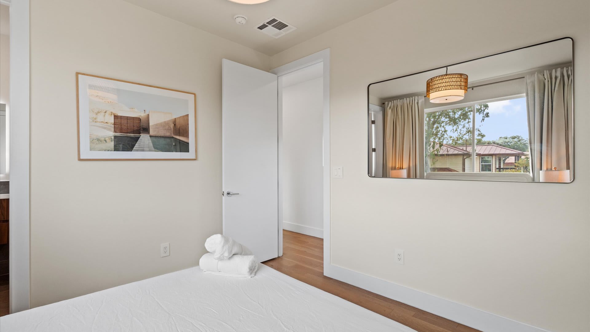 View of bedroom-2 showing a large rectangular mirror reflecting the window and bed, with a doorway leading to another room. The room has light walls and hardwood floors.