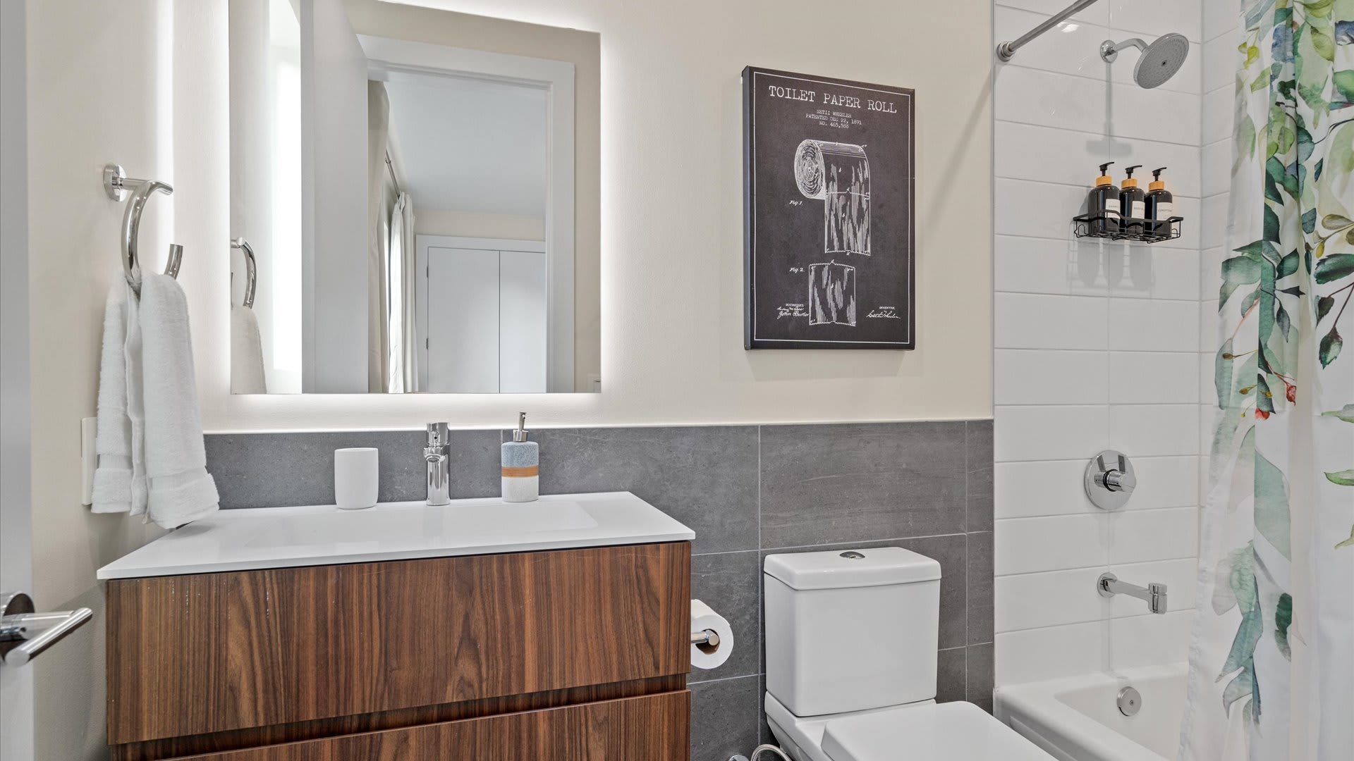 Bright bathroom with a white vanity, illuminated mirror, toilet, and a shower/tub combo with a leafy green shower curtain. Grey accent tiles line the lower wall.