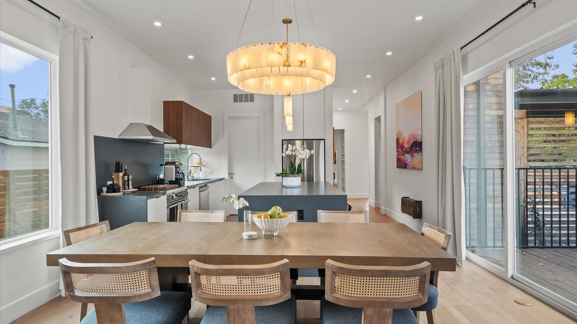 Bright dining area with a wooden table and cane-back chairs, illuminated by a large circular chandelier, seamlessly connecting to a modern kitchen. The kitchen features dark countertops, white cabinetry, and a sliding glass door leading to an outdoor area.