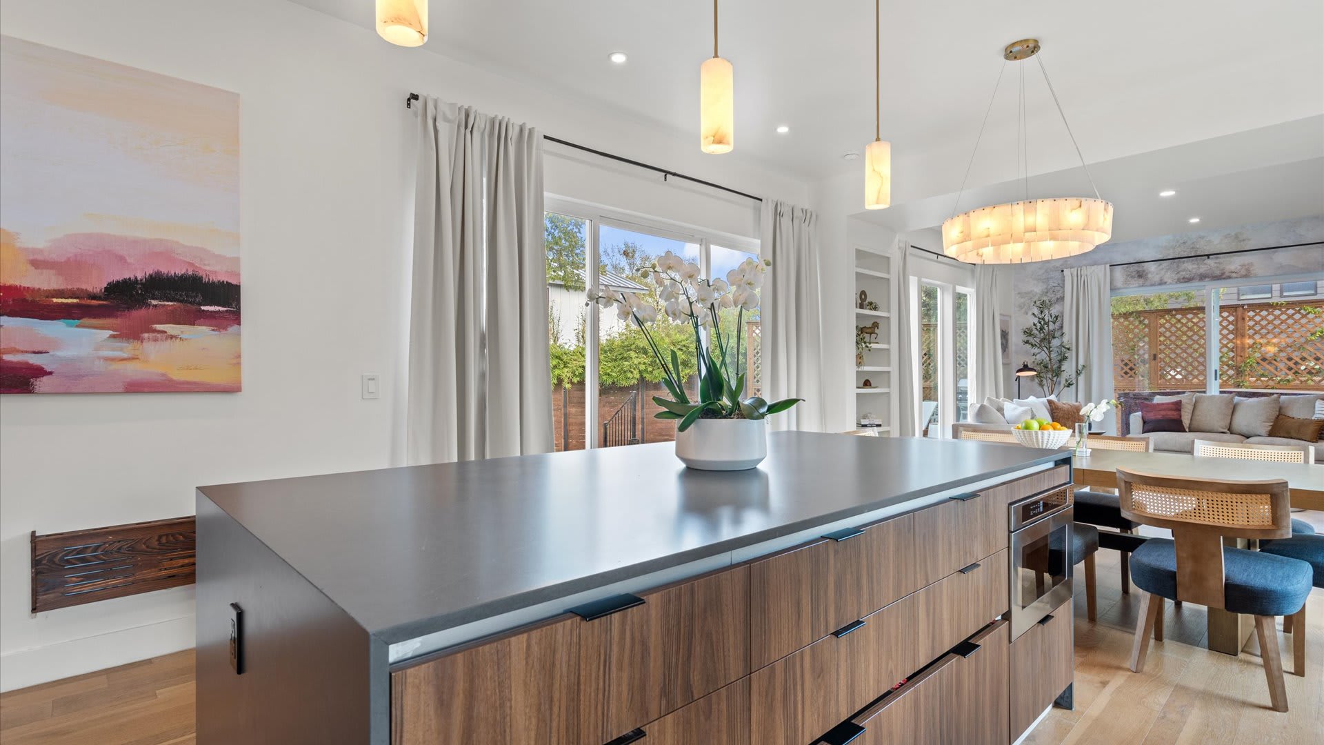 Bright open-concept kitchen with a large island featuring a dark countertop and rich wood-paneled drawers, adorned with an orchid centerpiece. The dining area and living room are visible in the background, illuminated by natural light.