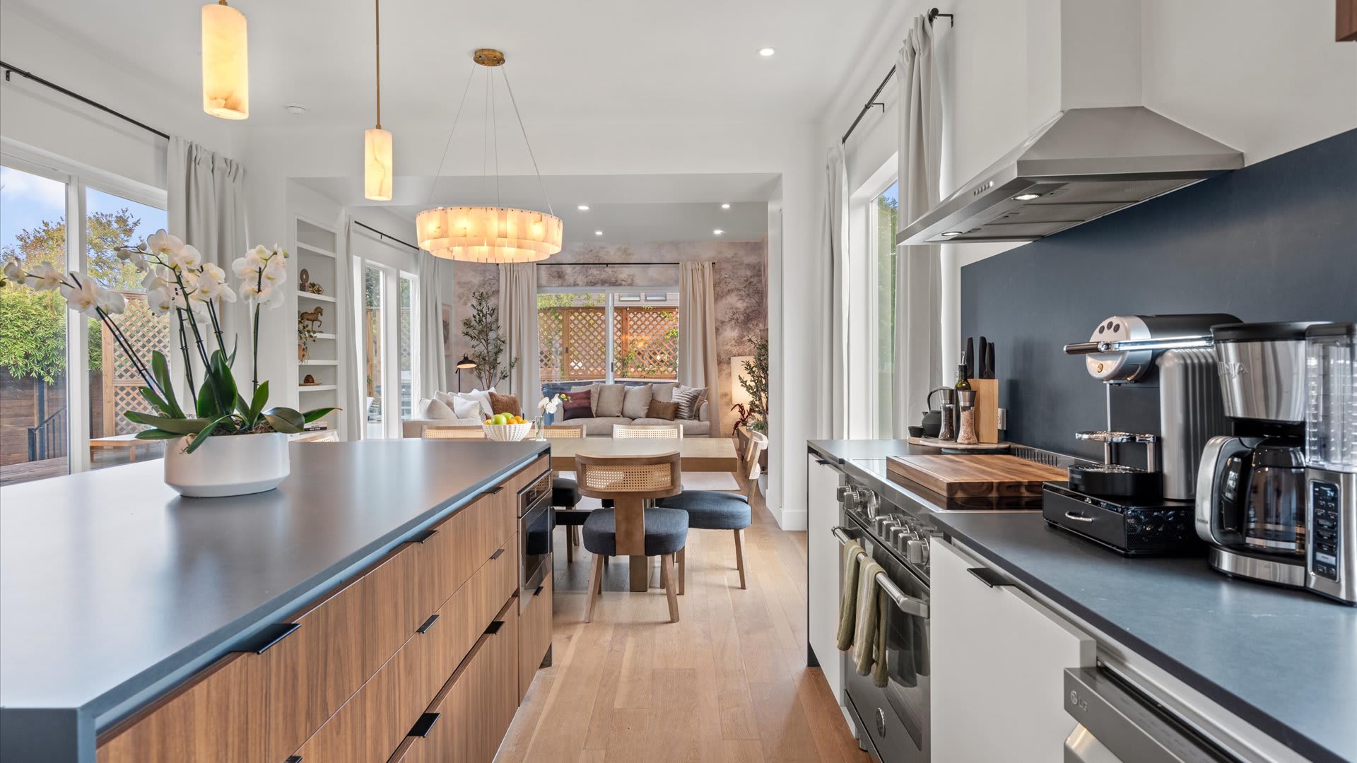 Contemporary kitchen with a long dark island, a gas range with a stainless steel hood, and a sink positioned under a window. The kitchen is well-equipped with a coffee maker and other small appliances, all set against light hardwood floors.
