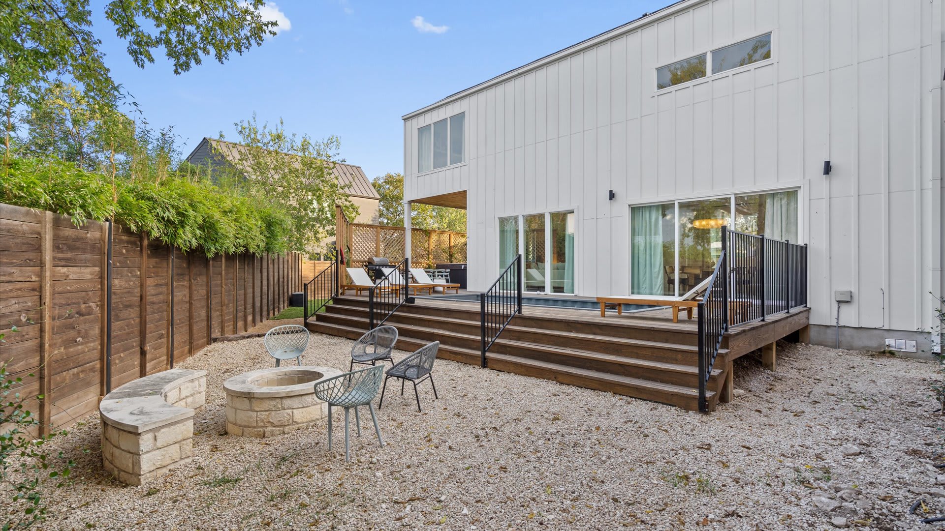 The backyard features a stone fire pit with built-in bench seating and modern wire chairs, situated on a gravel ground. A multi-level wooden deck with stairs leads up to the white modern house, offering distinct outdoor zones.
