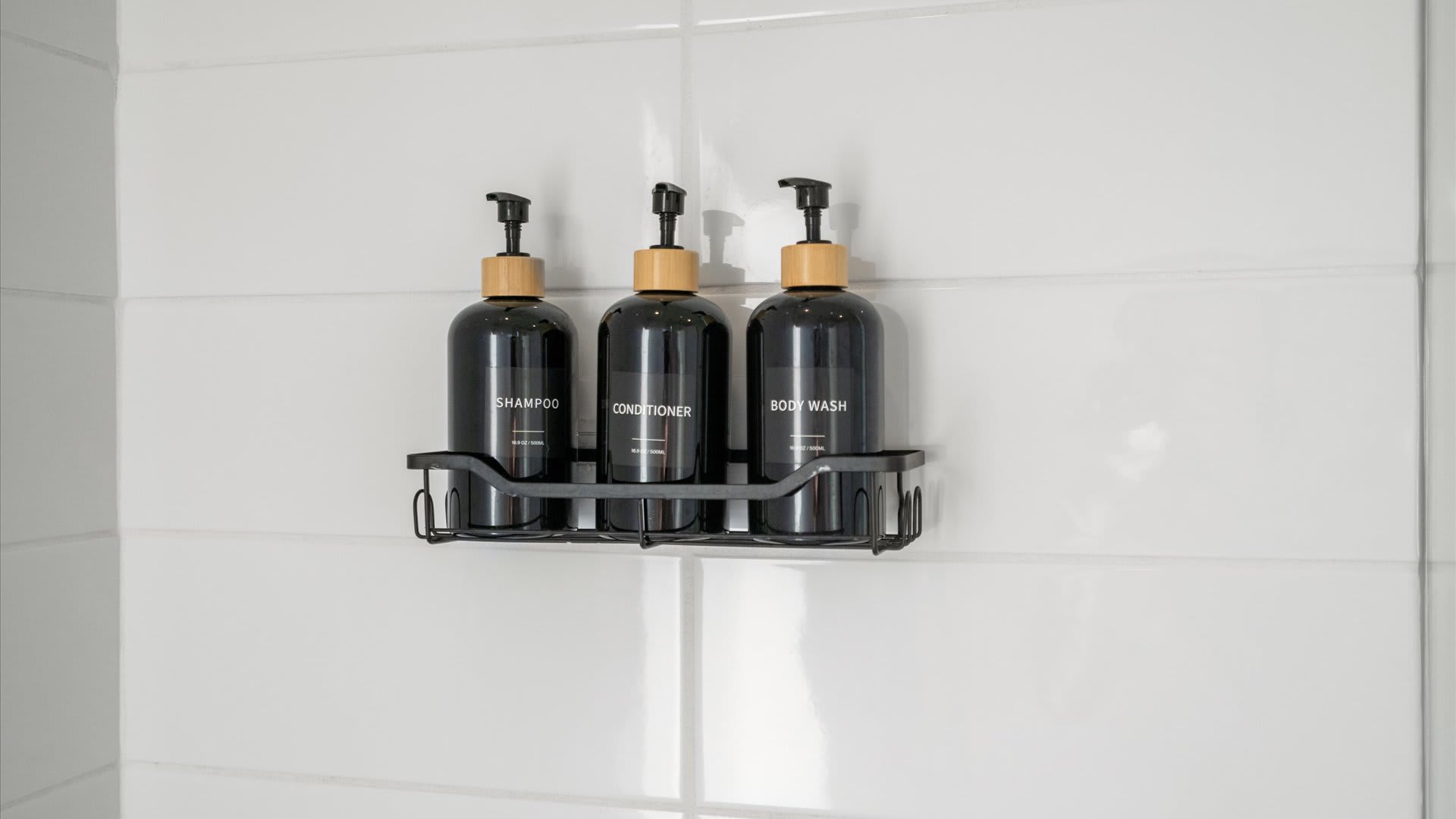 A close-up of three black pump bottles labeled "Shampoo," "Conditioner," and "Body Wash" in a black wire caddy, mounted on white subway tile in a shower.