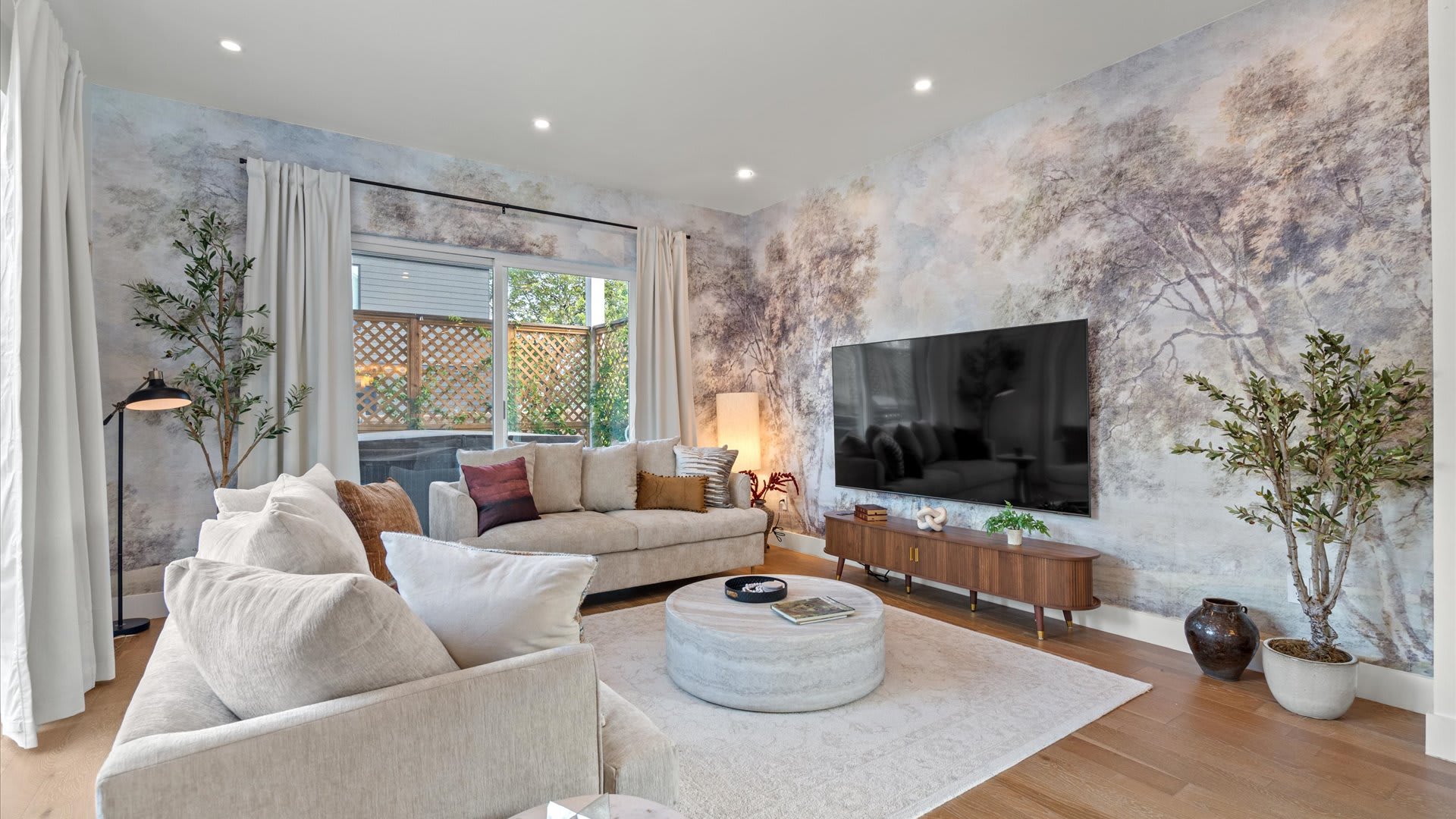 A cozy living room features a large wall covered in a scenic, muted tree mural wallpaper, with two light-colored sofas and a round ottoman on a patterned rug. A large flat-screen TV is mounted above a wooden media console, complemented by recessed lighting.
