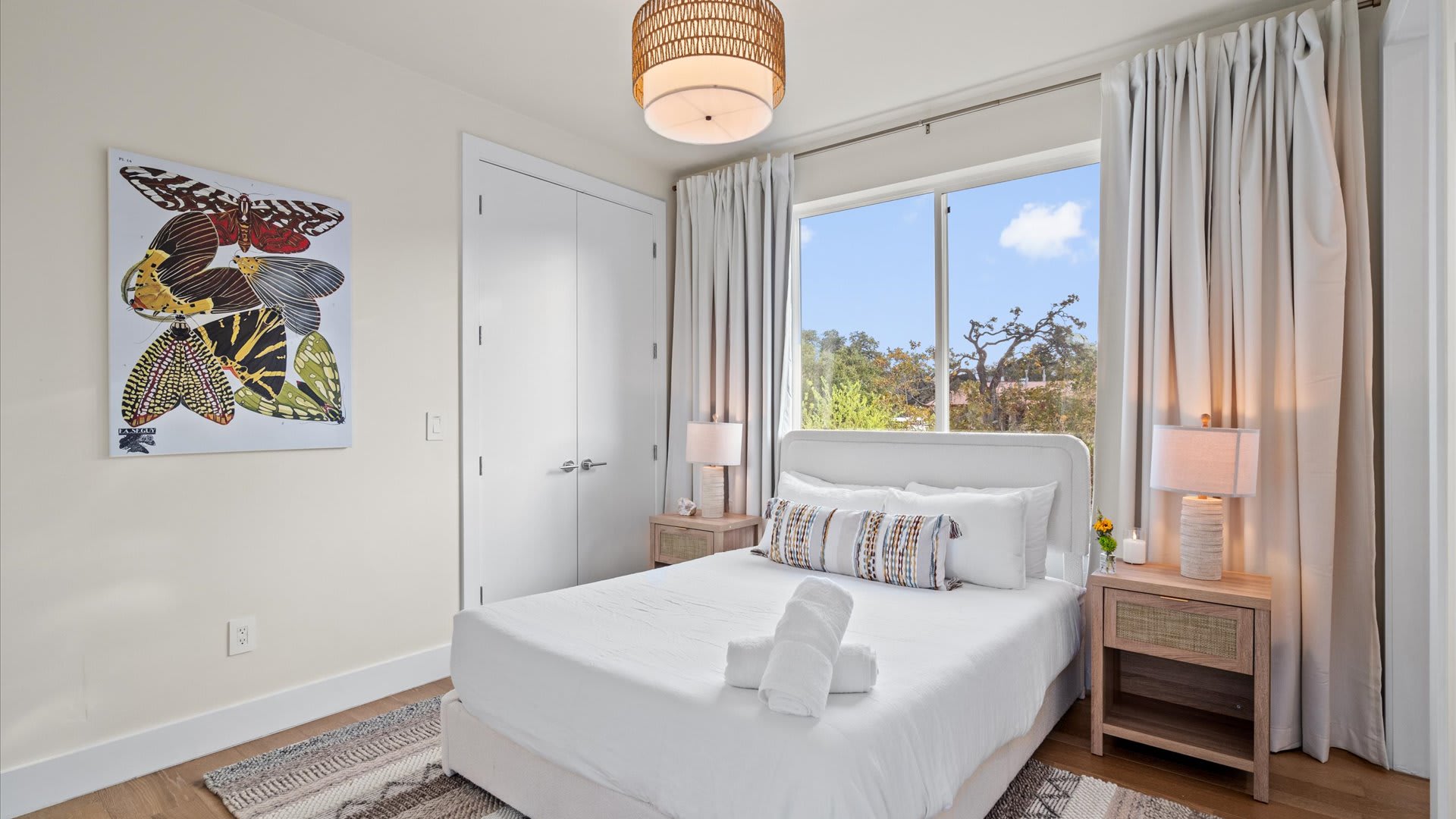 Cozy bedroom with a single bed, white linens, two wooden nightstands with lamps, and a large window with light-colored curtains overlooking trees. Butterfly wing artwork adorns one wall.
