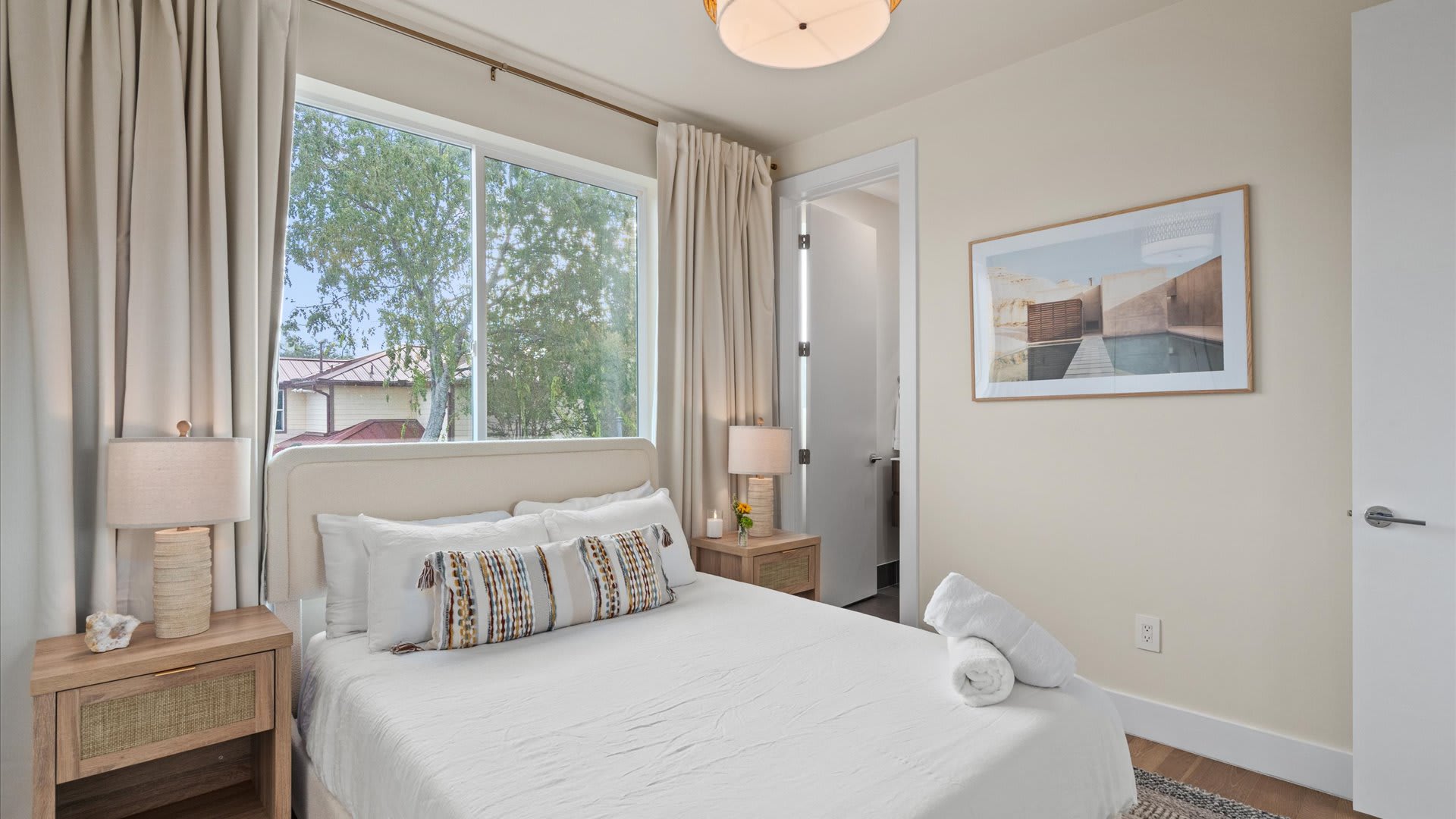 Close-up of bedroom-2's single bed with decorative pillows, flanked by wooden nightstands with lamps. A large window with light curtains provides natural light and a view of trees.