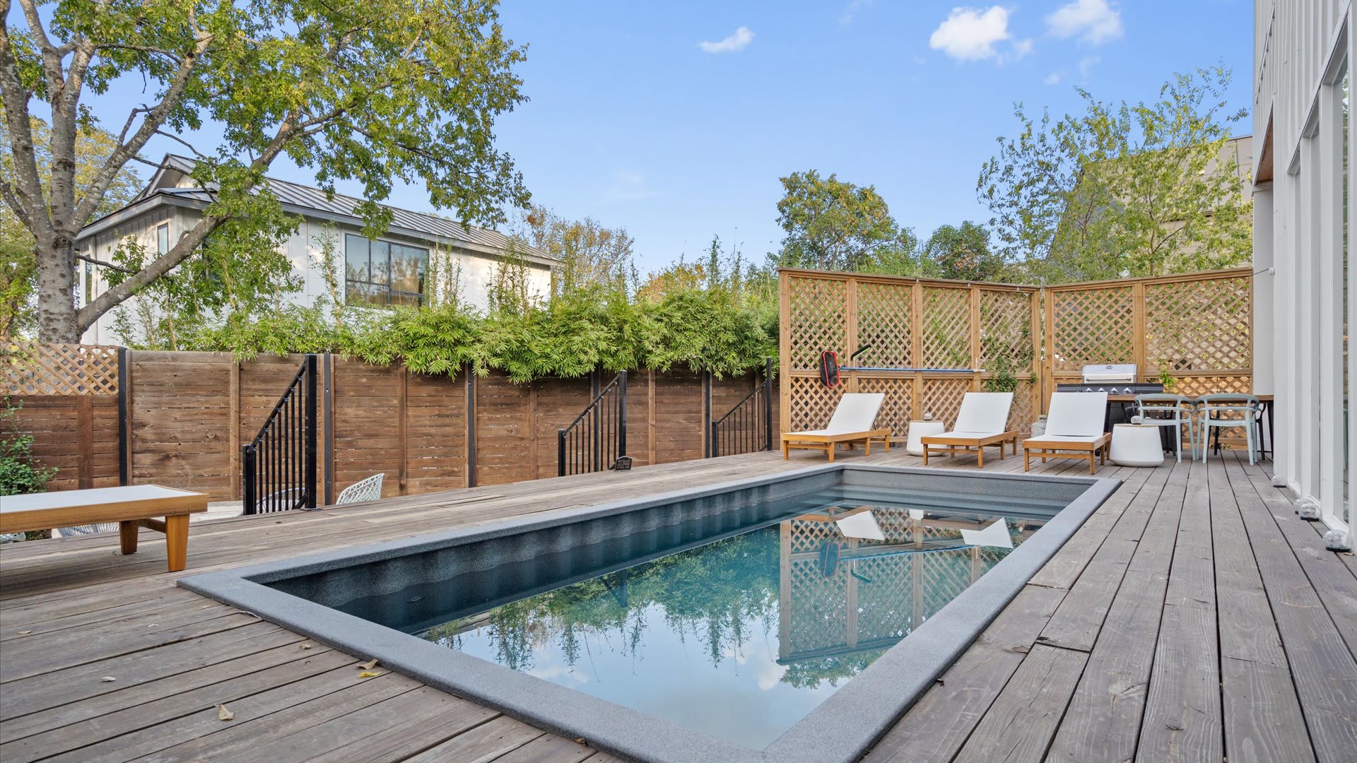 A ground-level view of the backyard shows a rectangular plunge pool integrated into a wooden deck, with lounge chairs and the modern white house in the background. A tall wooden fence provides privacy, creating a serene outdoor oasis.