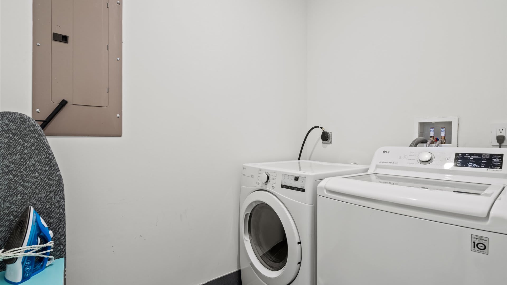A bright laundry room with white walls, featuring a white LG front-load washing machine and a top-load dryer, an ironing board, and an electrical panel on the wall.