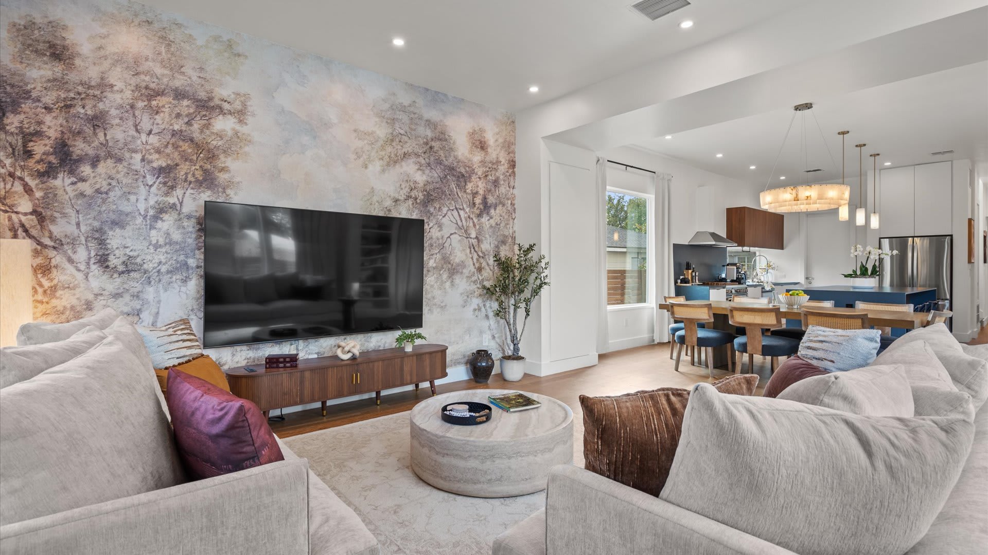 Open-concept living room featuring a large wall-mounted television above a wooden console, flanked by two light-colored sofas. A decorative mural wallpaper covers the accent wall, seamlessly transitioning into the dining and kitchen areas.